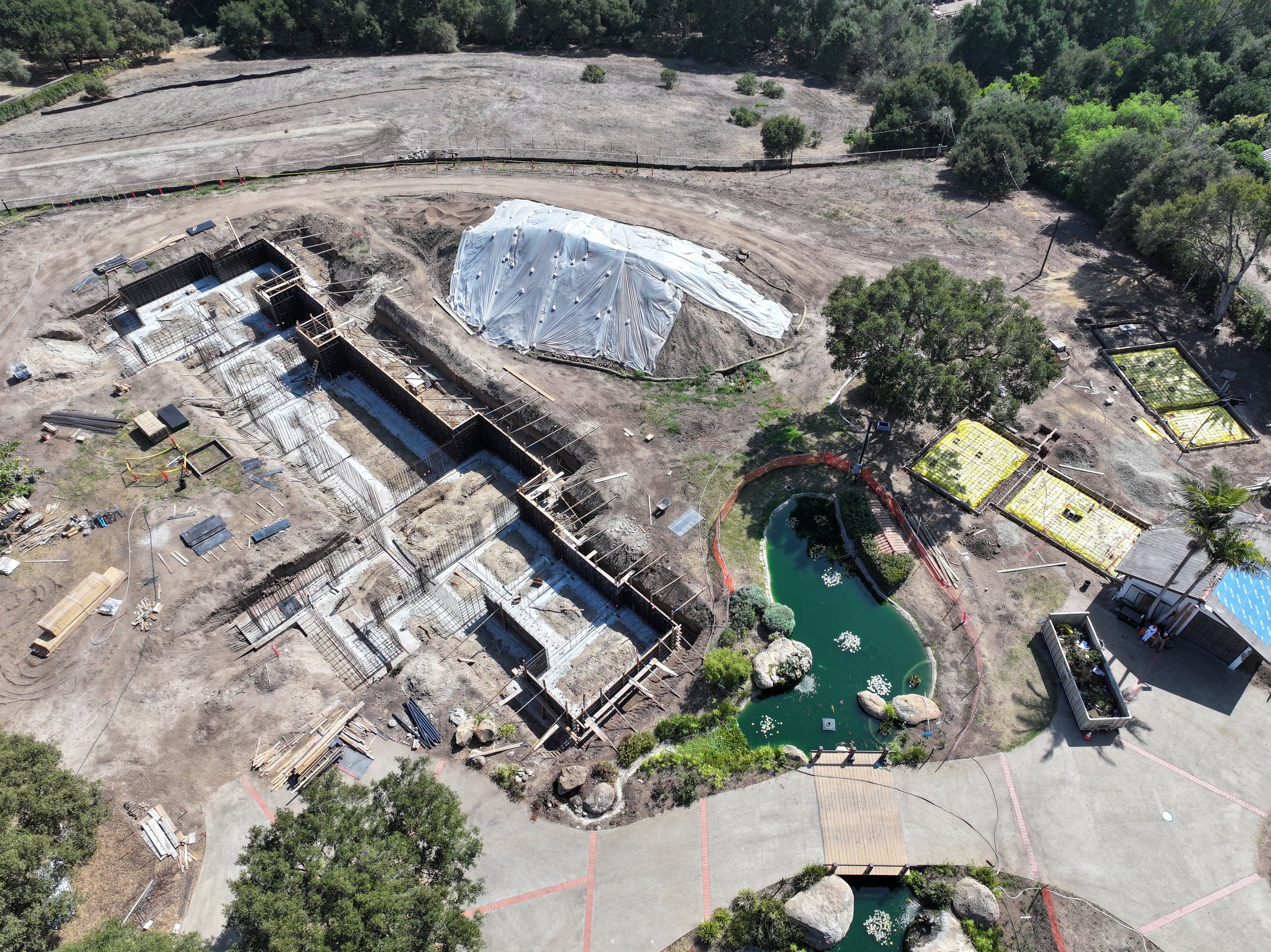 Aerial view of a construction site with foundation work, a covered structure, and a landscaped pond with large rocks and greenery.