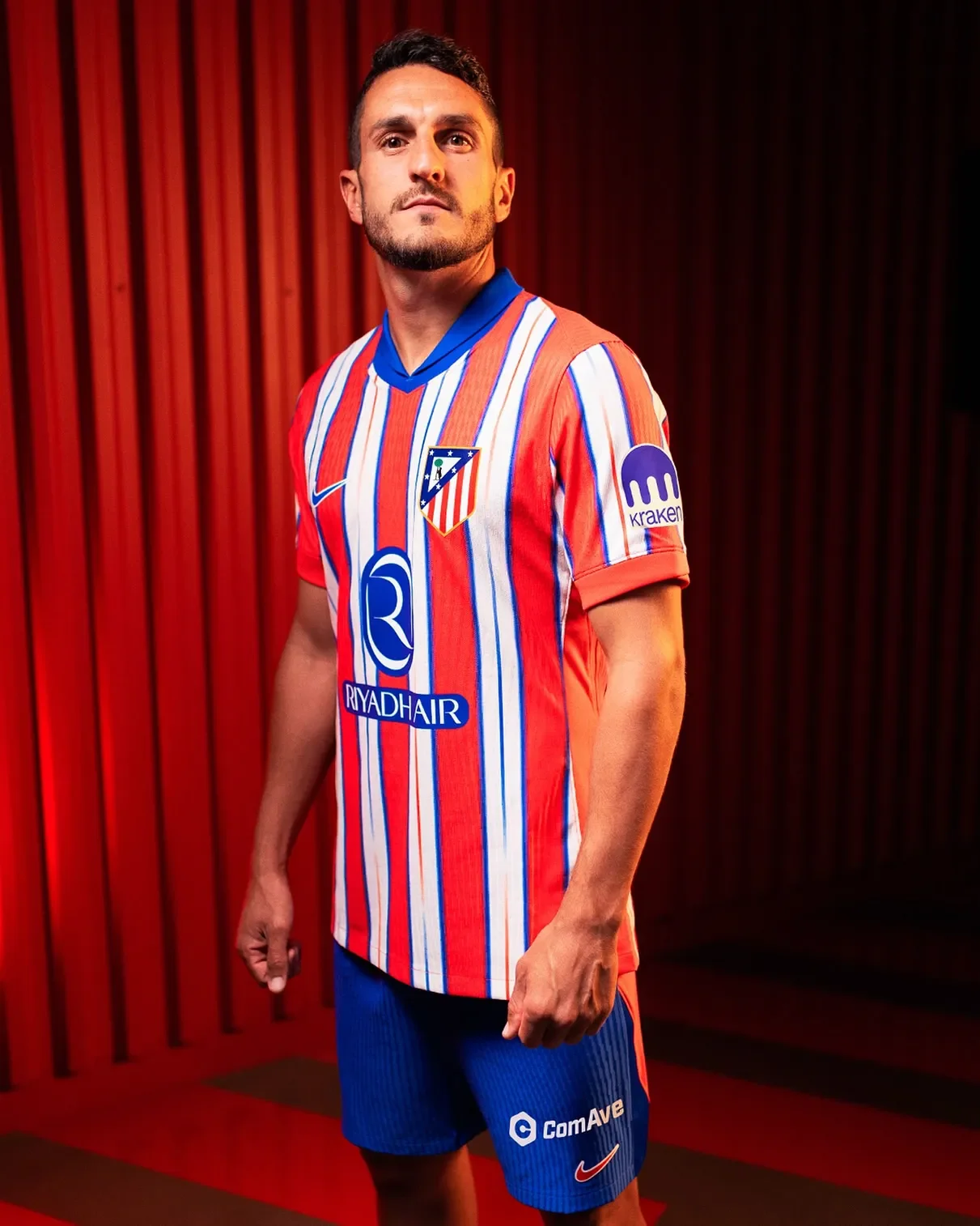 Hombre con uniforme del Atlético de Madrid, camiseta a rayas rojas, blancas y azules, con patrocinadores en el frente. Fondo de madera roja.