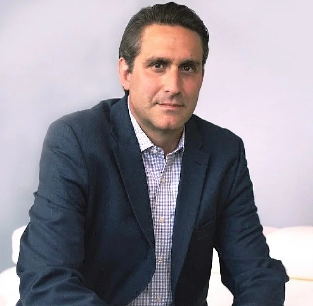 A man with dark hair in a combed style, wearing a navy blazer over a checkered shirt, sitting against a light gray background.