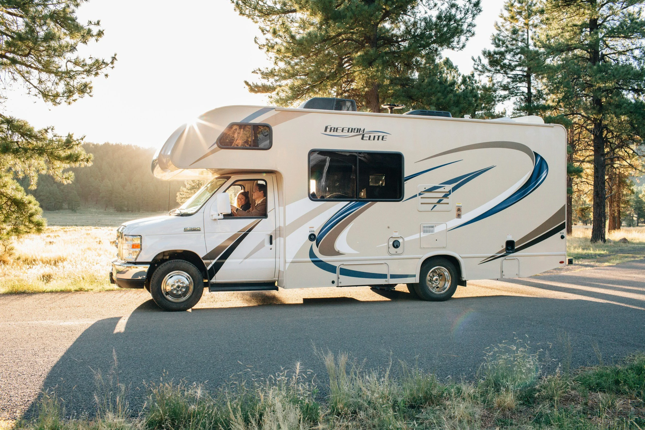 A white motorhome with blue and beige decals parked on a paved road in a forested area during sunset, with two people inside.