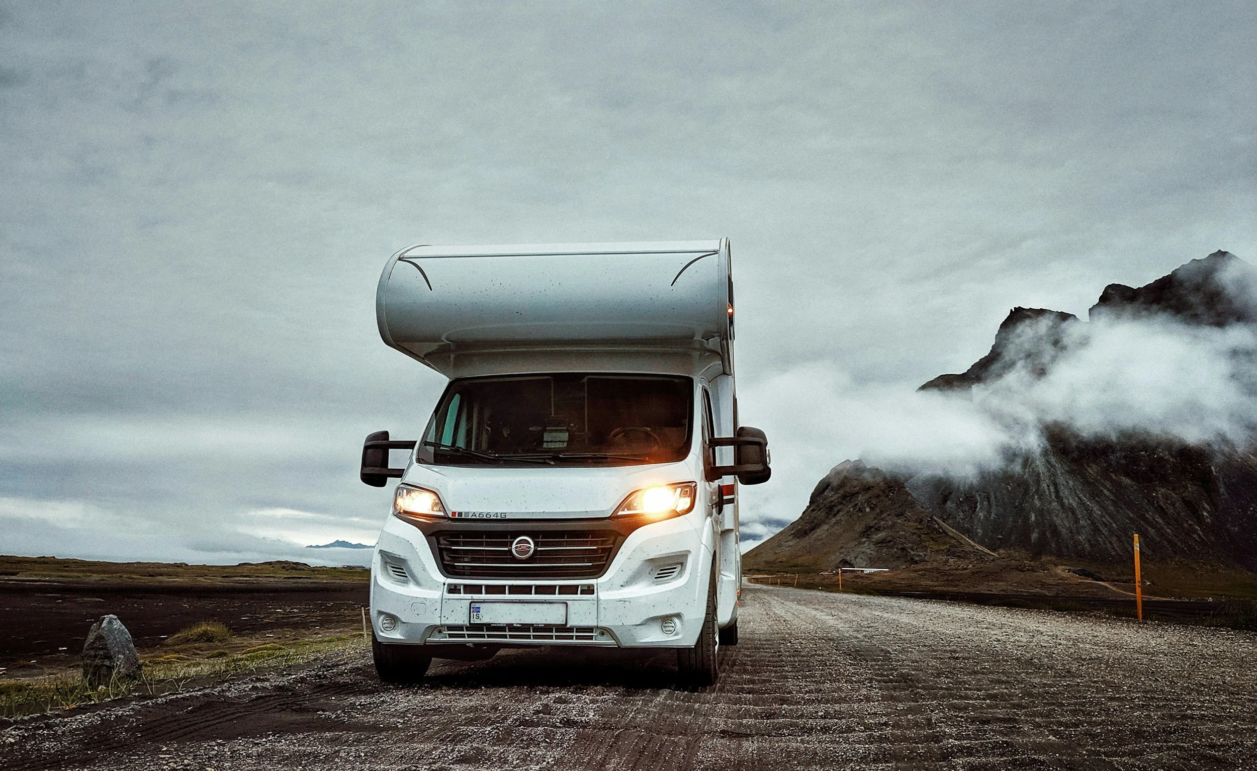 White camper van traveling on a gravel road with mountainous landscape and cloudy sky in the background.