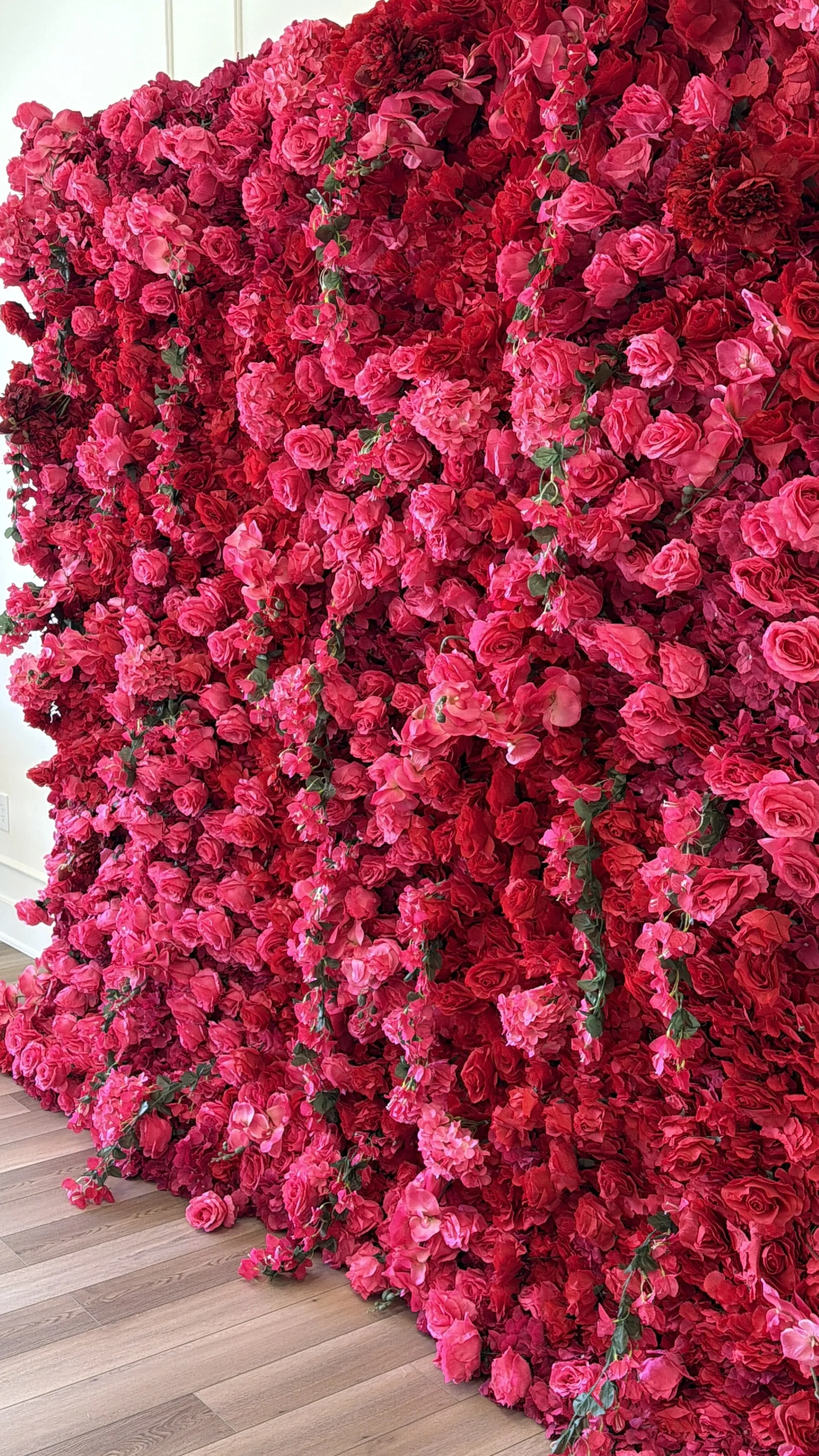 wall covered with a large display of pink and red artificial flowers, with some green leaves, on a wooden floor and white wall background.