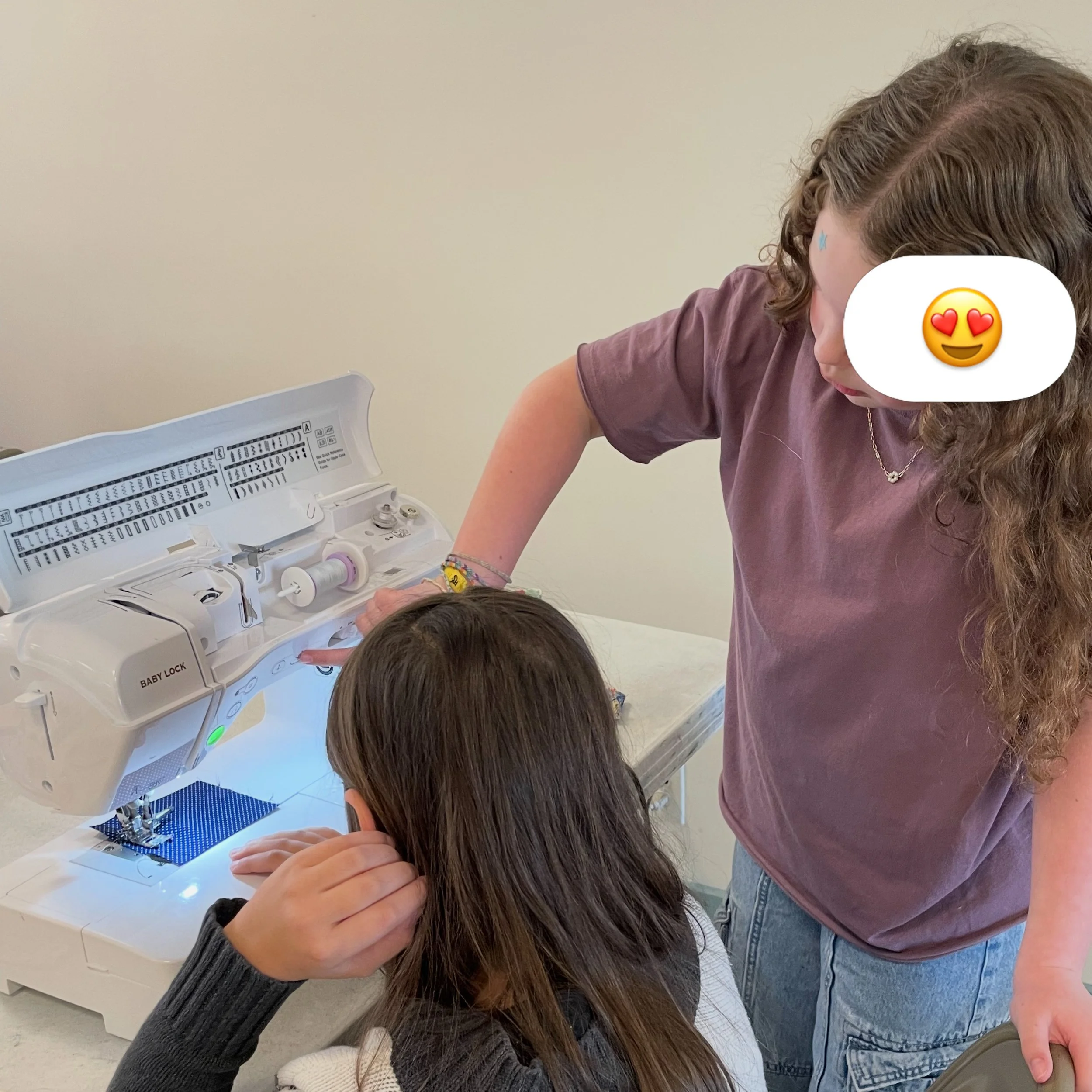 A young girl with long brown hair is sitting at a sewing machine, sewing fabric. She is wearing a black long-sleeved shirt and beige pants. The setting appears to be a light-colored room with minimal background details.