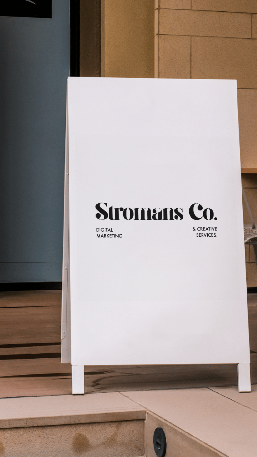 White sandwich board sign with black text reading "Stromans Co. DIGITAL MARKETING & CREATIVE SERVICES" positioned on a sidewalk outside a building.