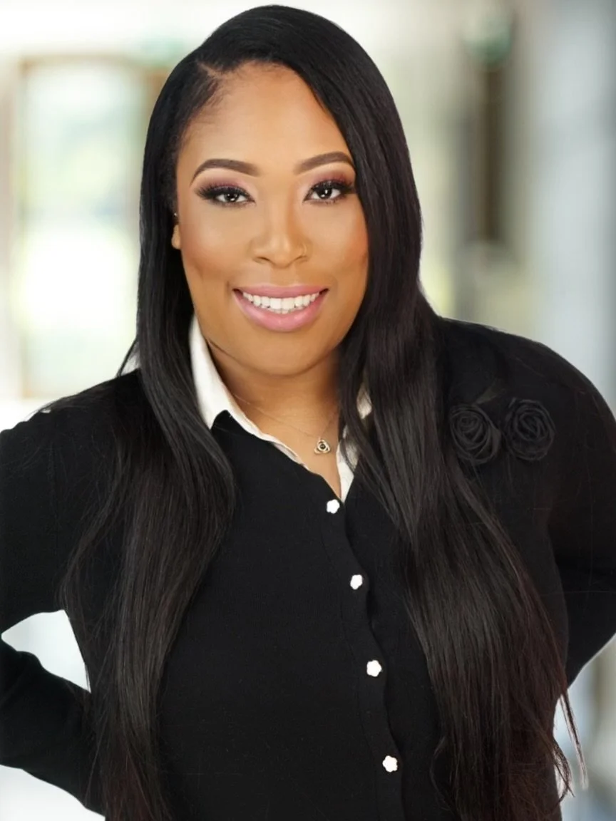 A woman with long black hair smiling, wearing a black button-up top with a white collar, standing in a well-lit indoor space.