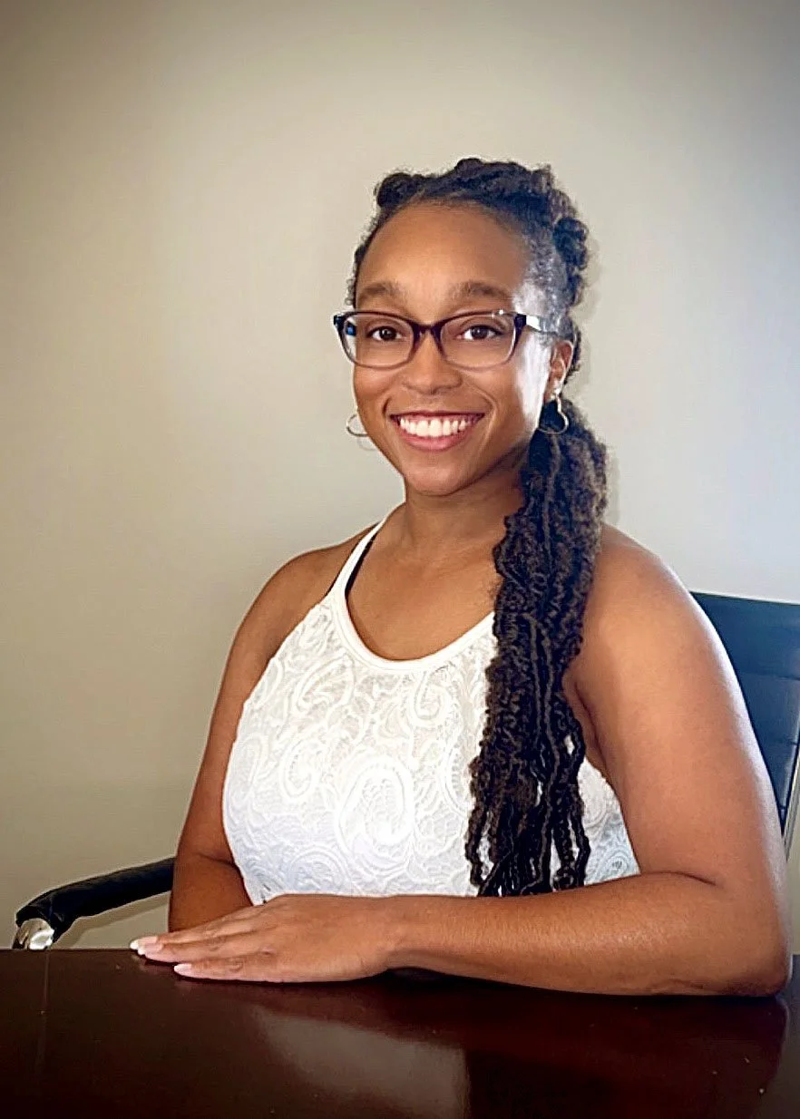 A woman with glasses, long curly hair, and hoop earrings, sitting at a desk in a white sleeveless top, smiling at the camera.