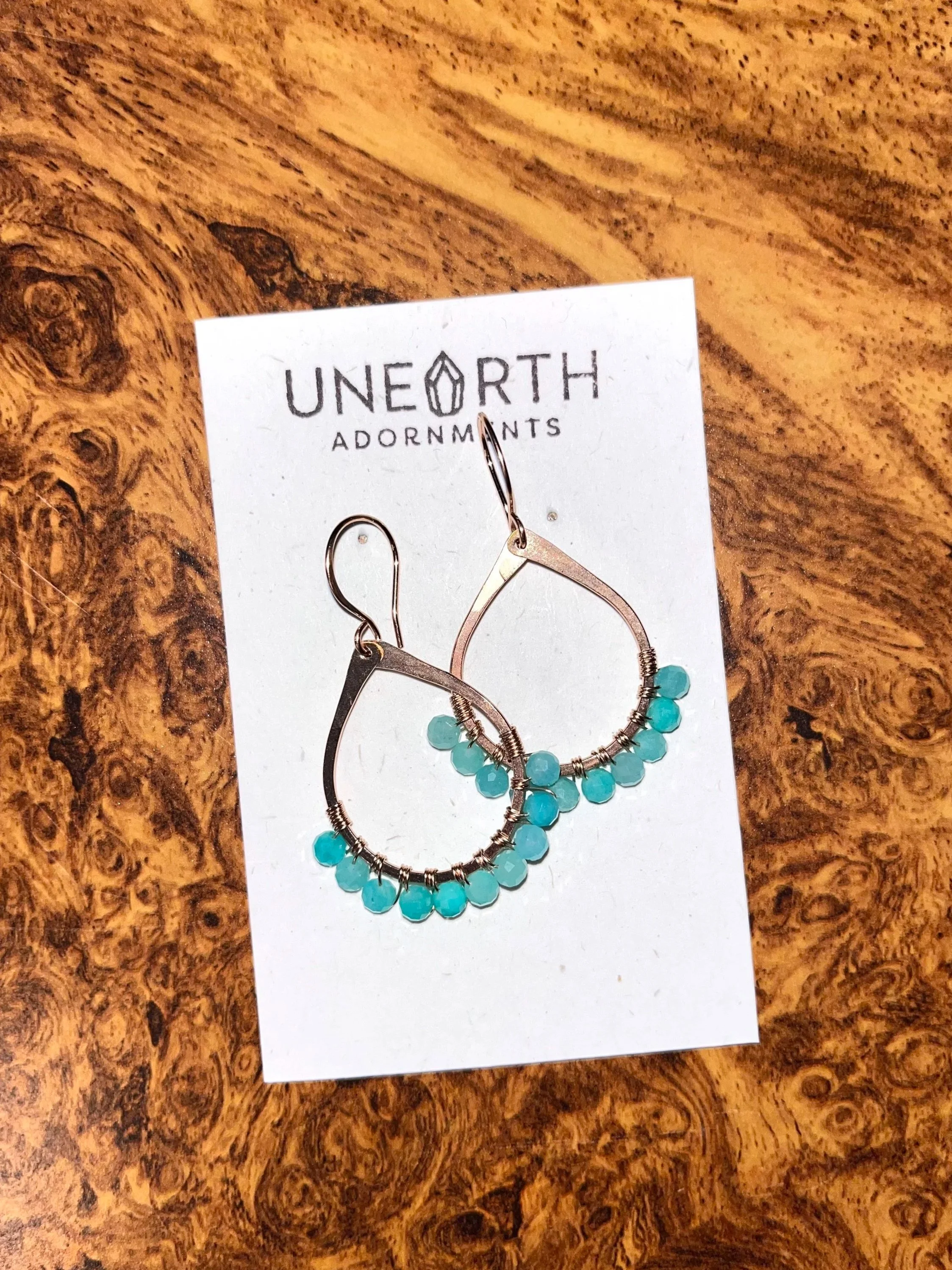 Pair of silver hoop earrings with turquoise beads on white card labeled 'Unearth Adornments' placed on a wooden surface.