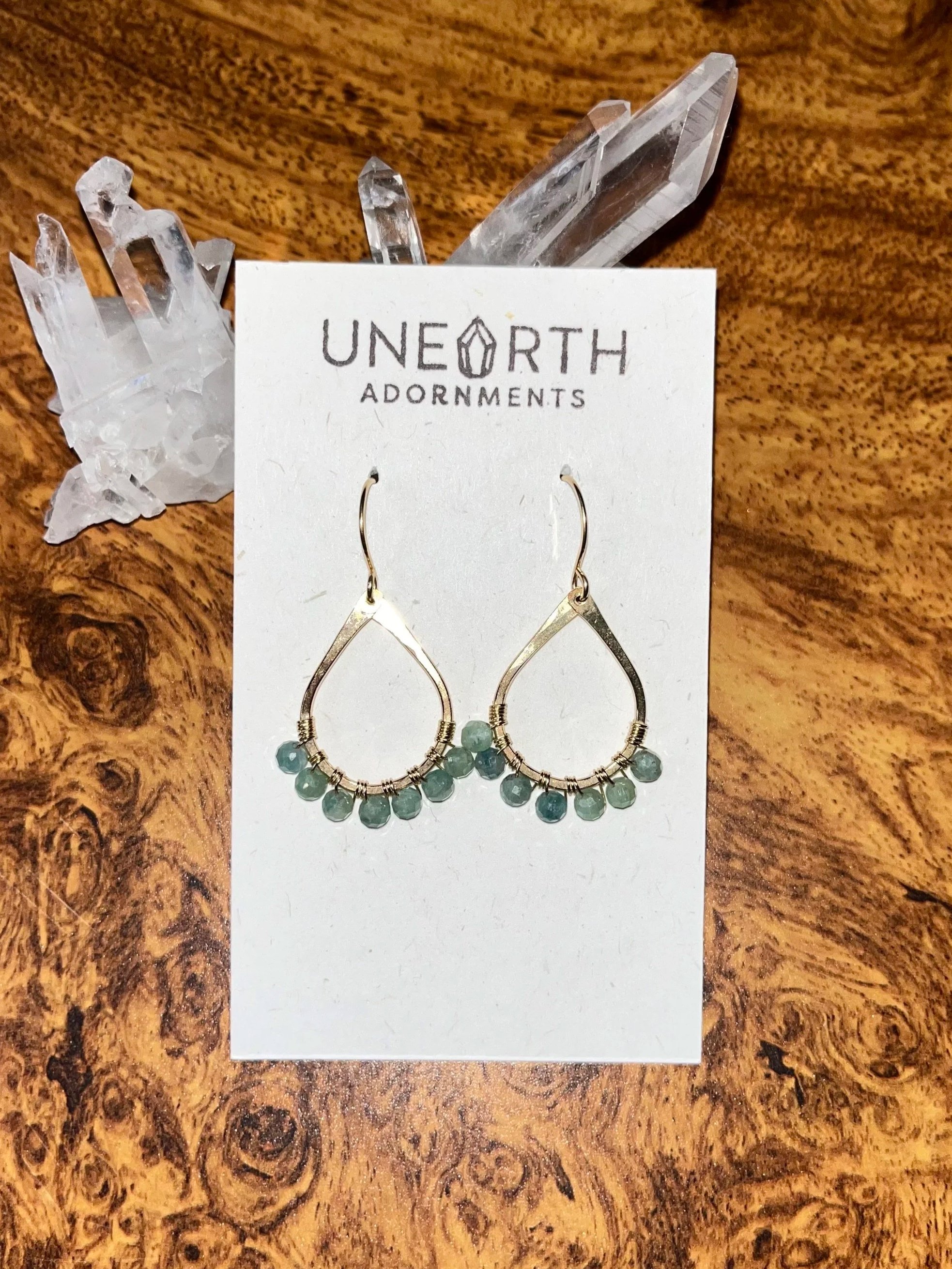 Pair of gold-filled earrings with green kyanite beads on a white jewelry display card labeled 'Unearth Adornments', placed on a wooden surface, with clear quartz crystals in the background.