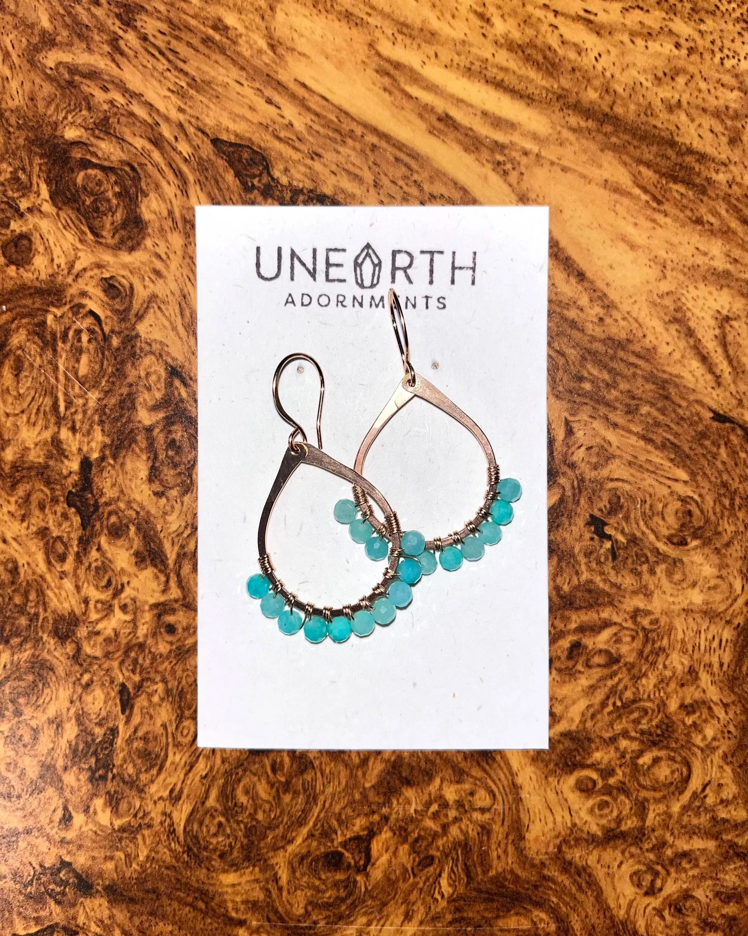 Rose gold teardrop-shaped earrings with amazonite gemstones hanging from a white card on a wooden surface.