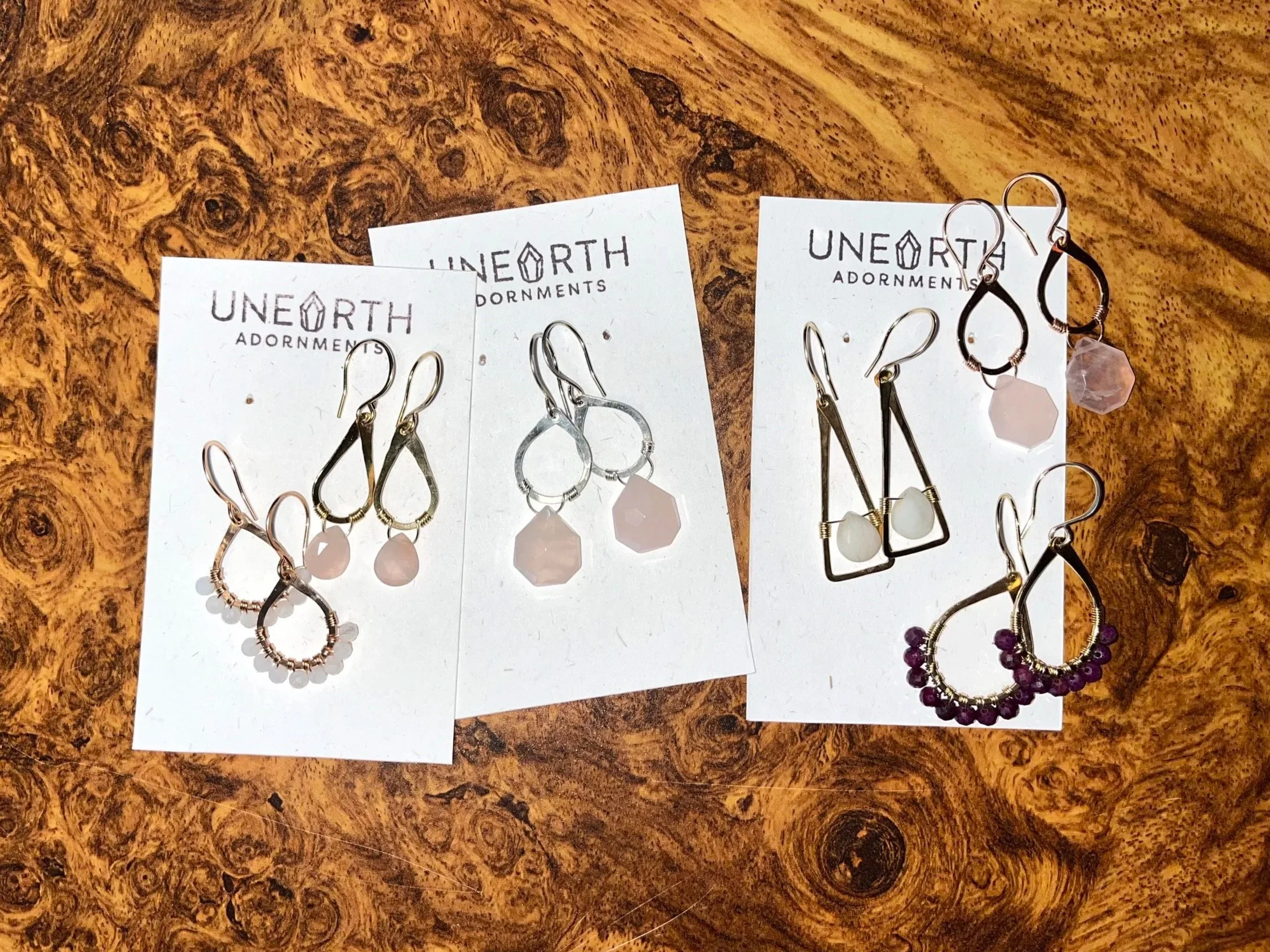 Display of five pairs of earrings on three white cards on a wooden surface. The earrings are of various styles and colors, including rose gold, silver, black, and gold, with some featuring rose quartz, ruby, moonstone gemstones