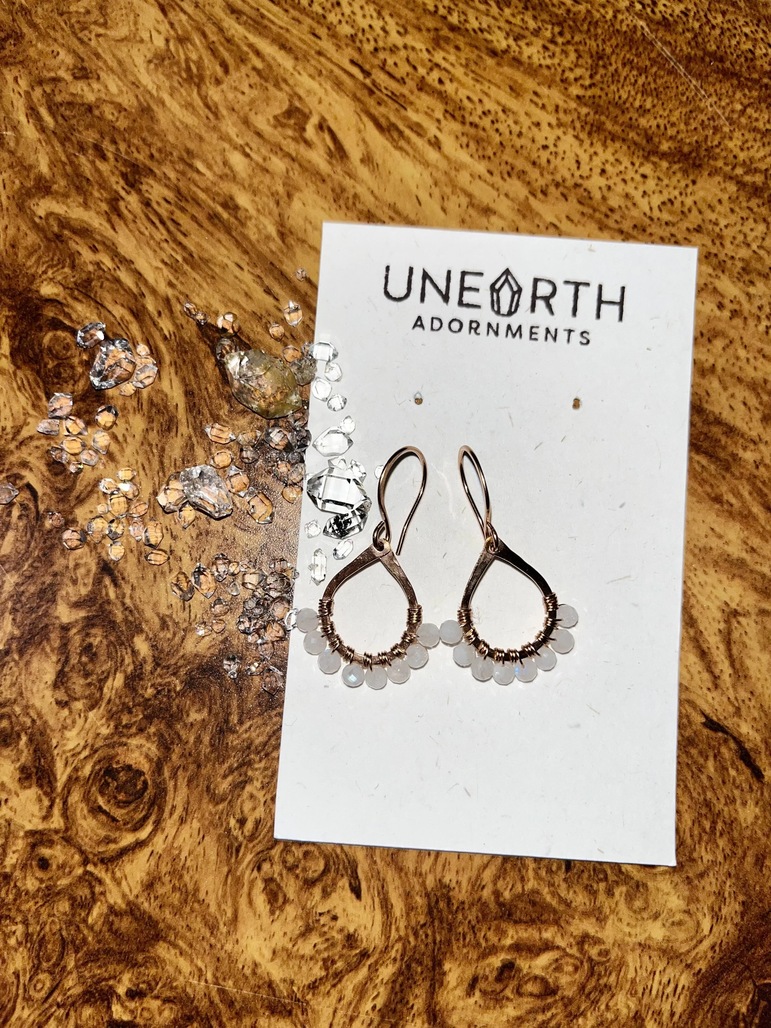Rose gold earrings with moonstone beads on white card labeled UNEARTH ADORNMENTS, placed on a wooden surface with scattered small clear rhinestones.