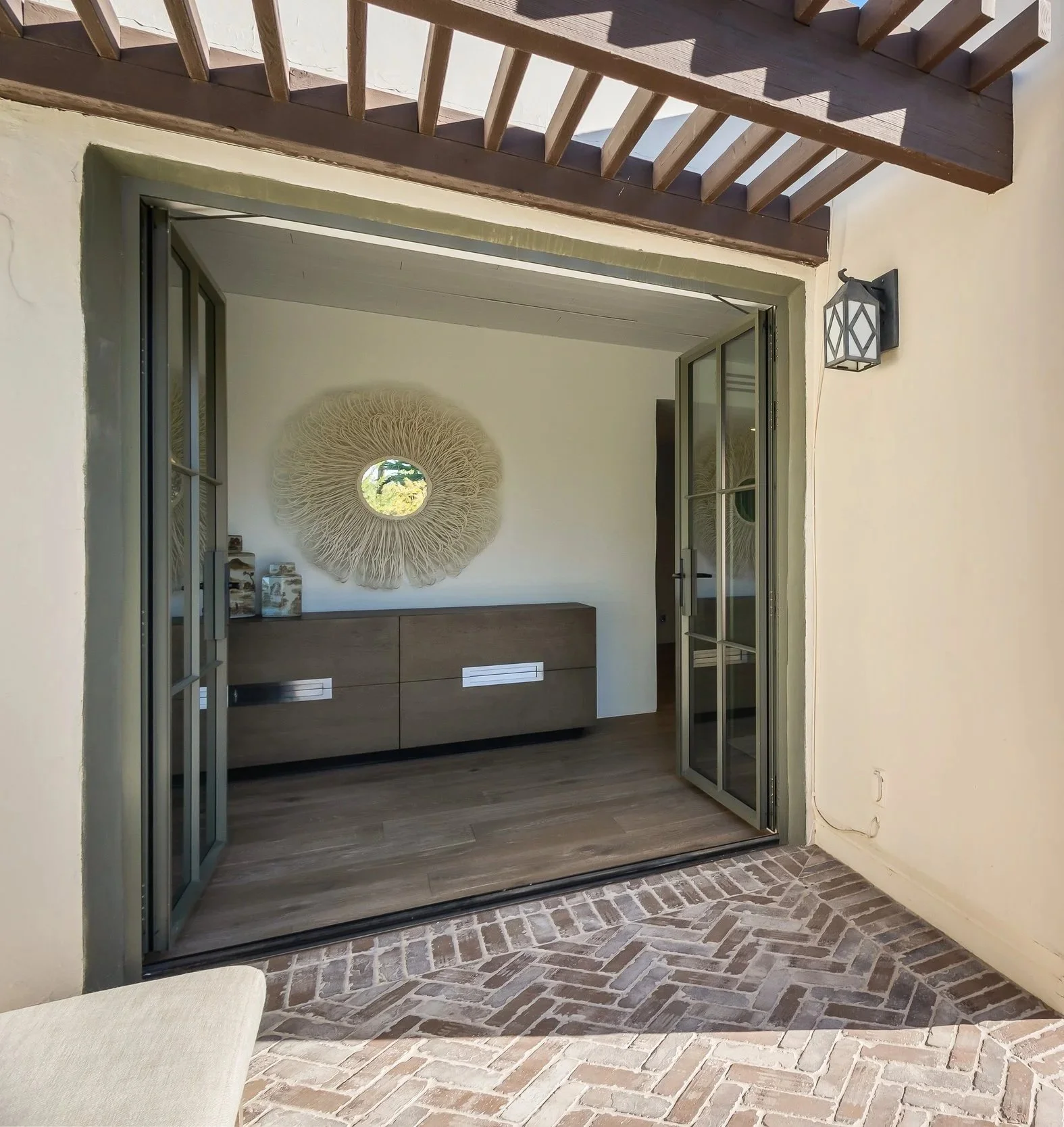 View of an open sliding glass door leading into a room with a wall decor mirror, a wooden sideboard, and decorative items inside.