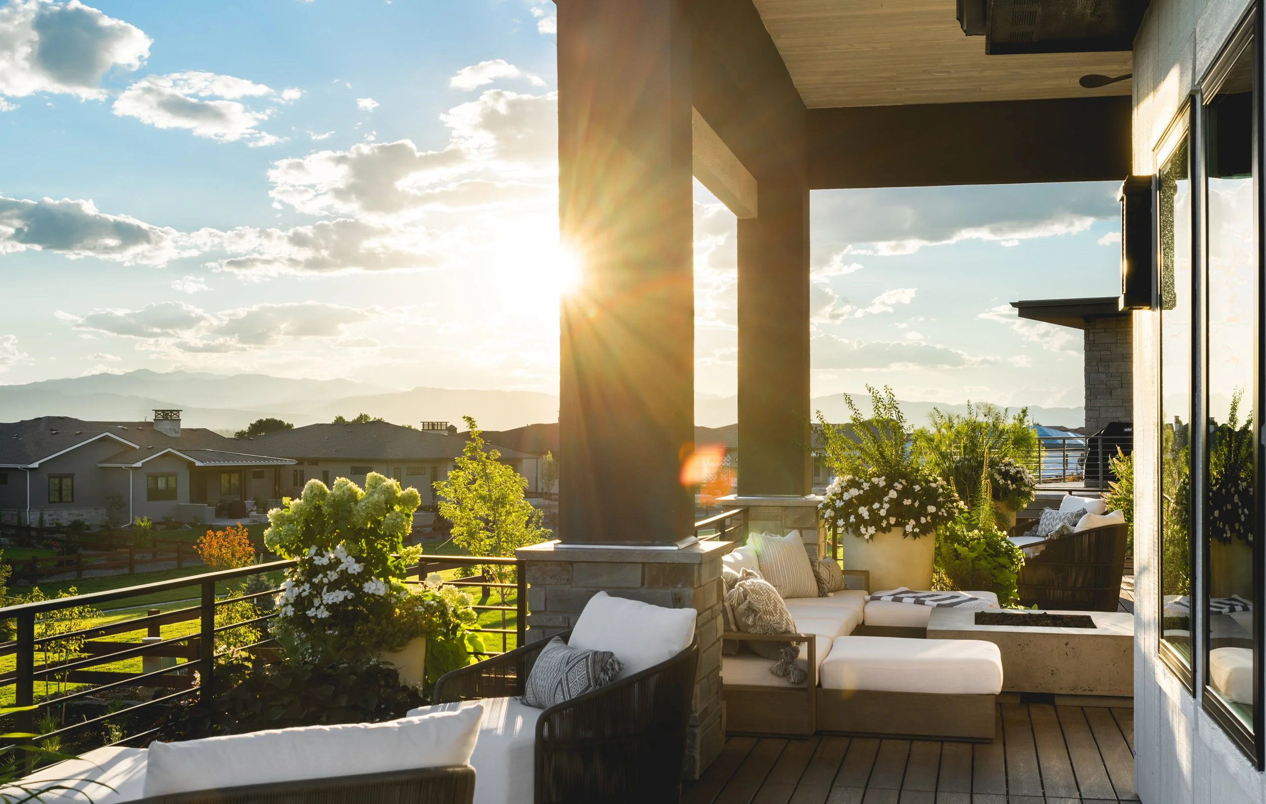 An outdoor balcony or patio area with seating and potted plants, overlooking a suburban neighborhood with houses, trees, and mountains in the distance, during sunset.