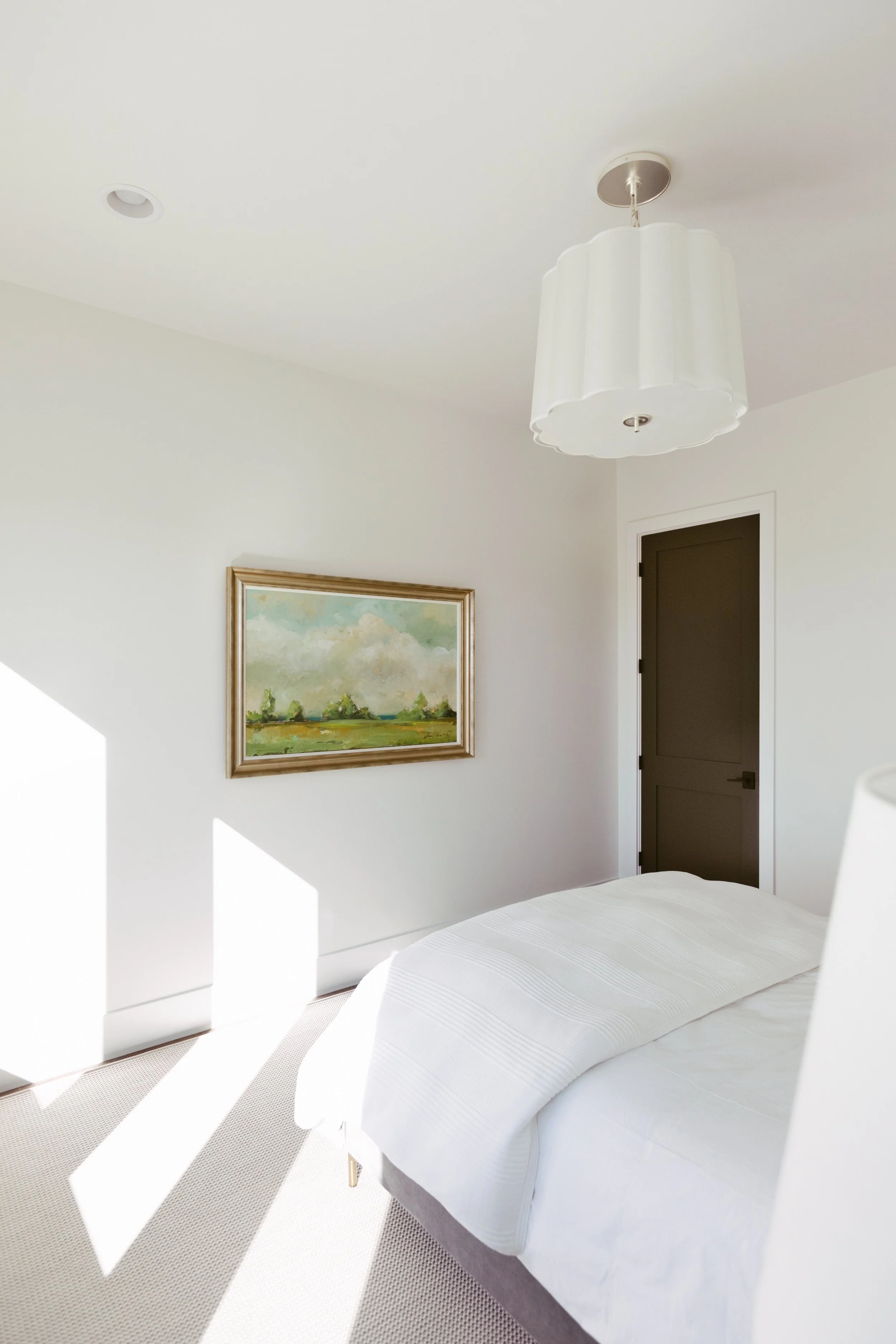 Bright bedroom with sunlight streaming through a window, a white bed, a framed landscape painting on the wall, a white ceiling light fixture, and a dark-colored door.