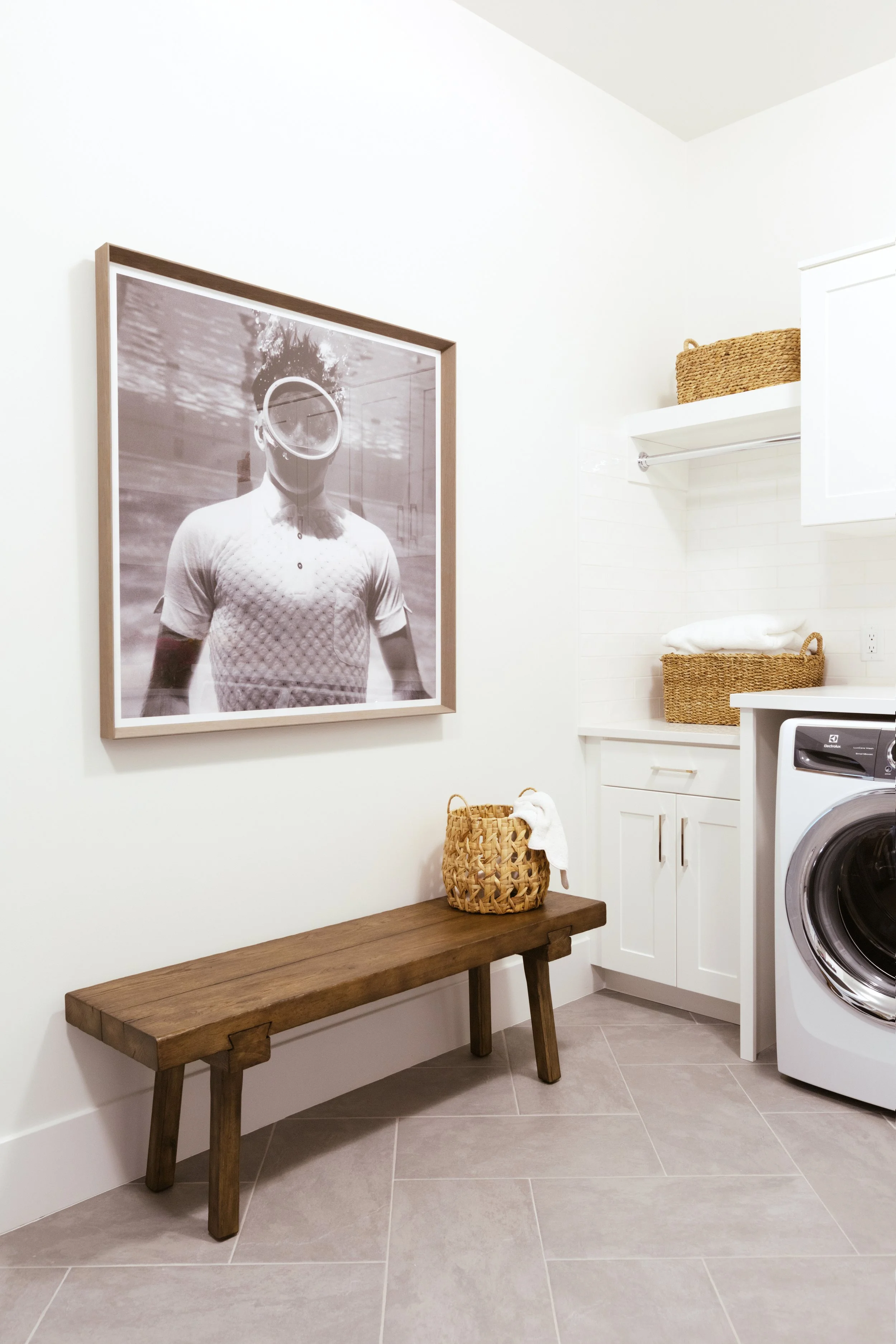 A laundry room with a framed black-and-white photo of a person wearing goggles. The room has white walls, a wooden bench, wicker baskets, white cabinets, and a front-loading washing machine.