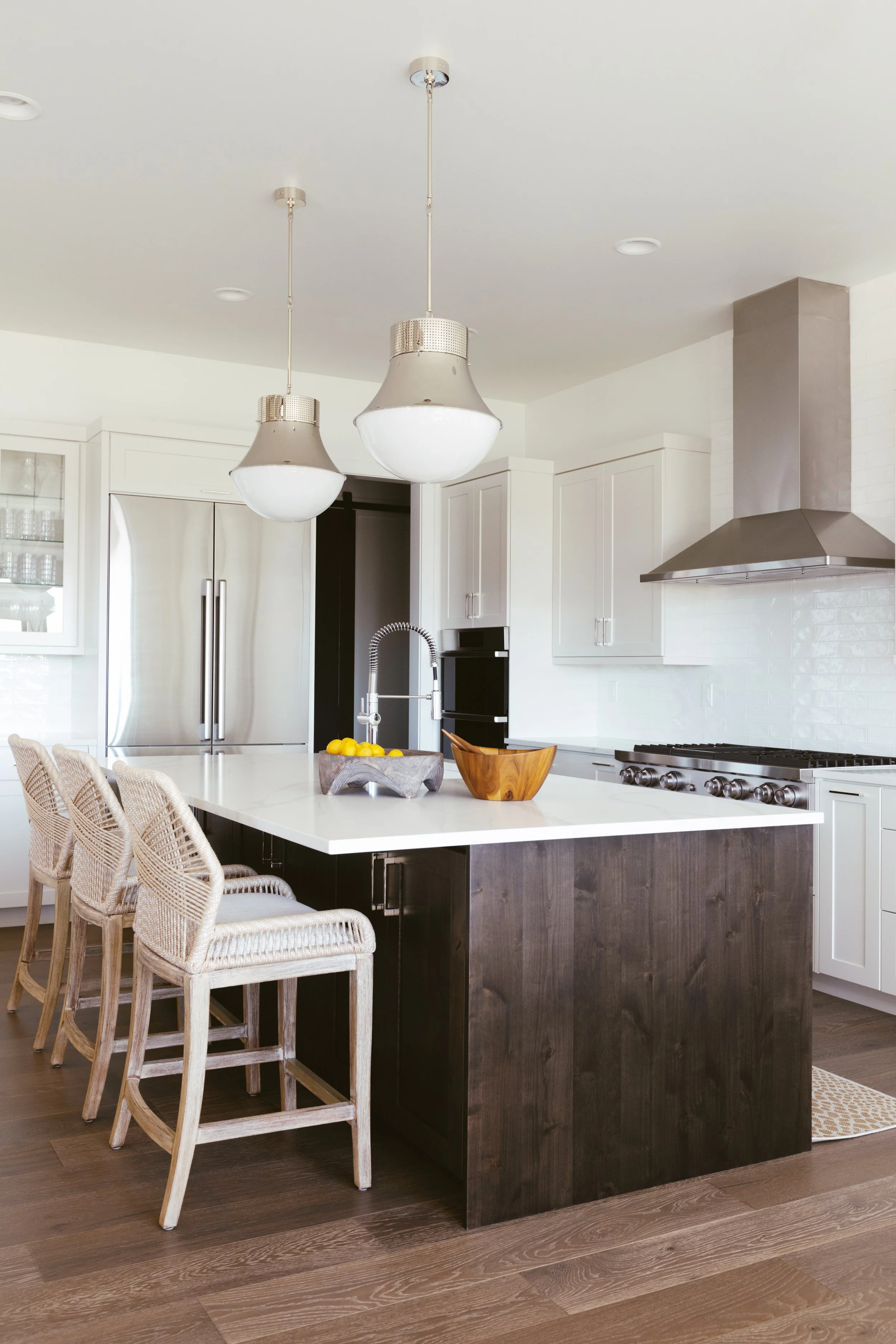 Modern kitchen with white cabinets, a large island with a white countertop and dark wooden base, three woven barstools, stainless steel refrigerator, pendant lights, built-in oven, stove, and stainless steel range hood.