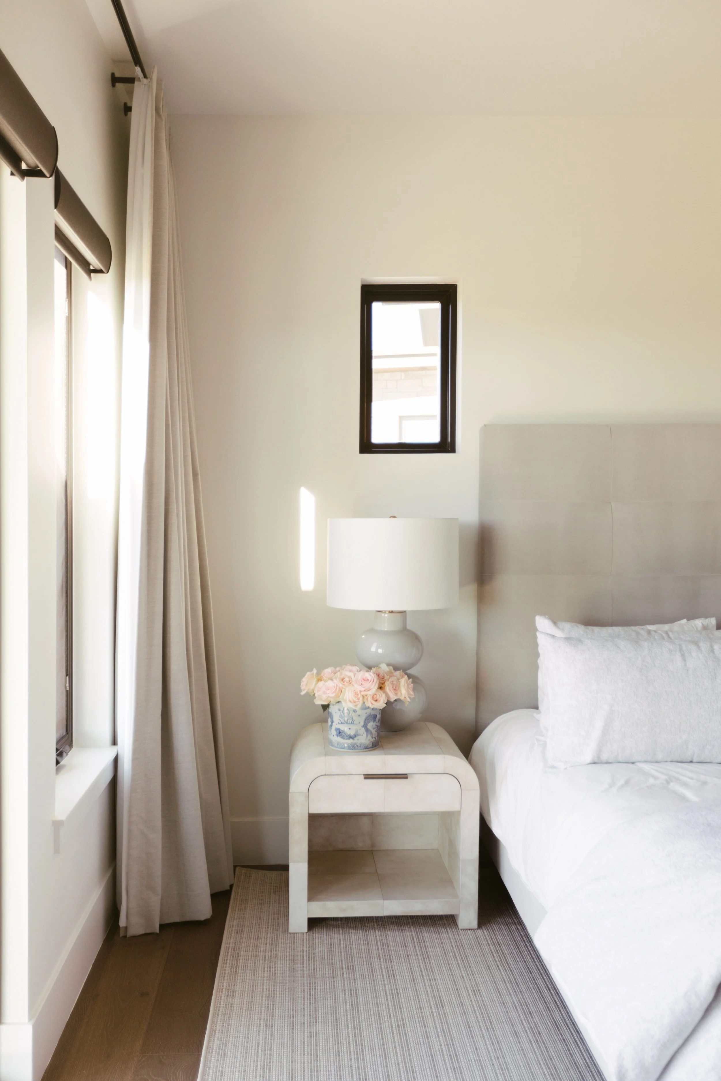 A bedroom corner with a bed, white pillows, a bedside table, a lamp, a vase with pink roses, a window, and beige curtains.