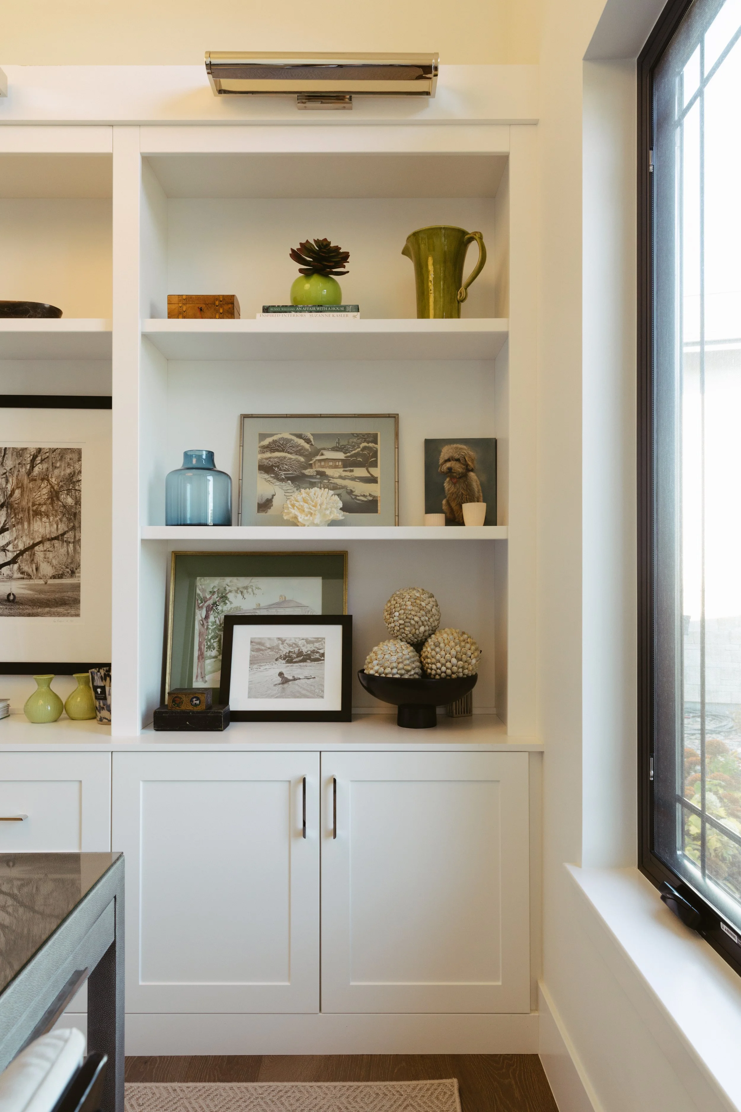 Interior view of a white built-in cabinet with decorative items, framed photos, vases, and a bowl of decorative balls, next to a window.