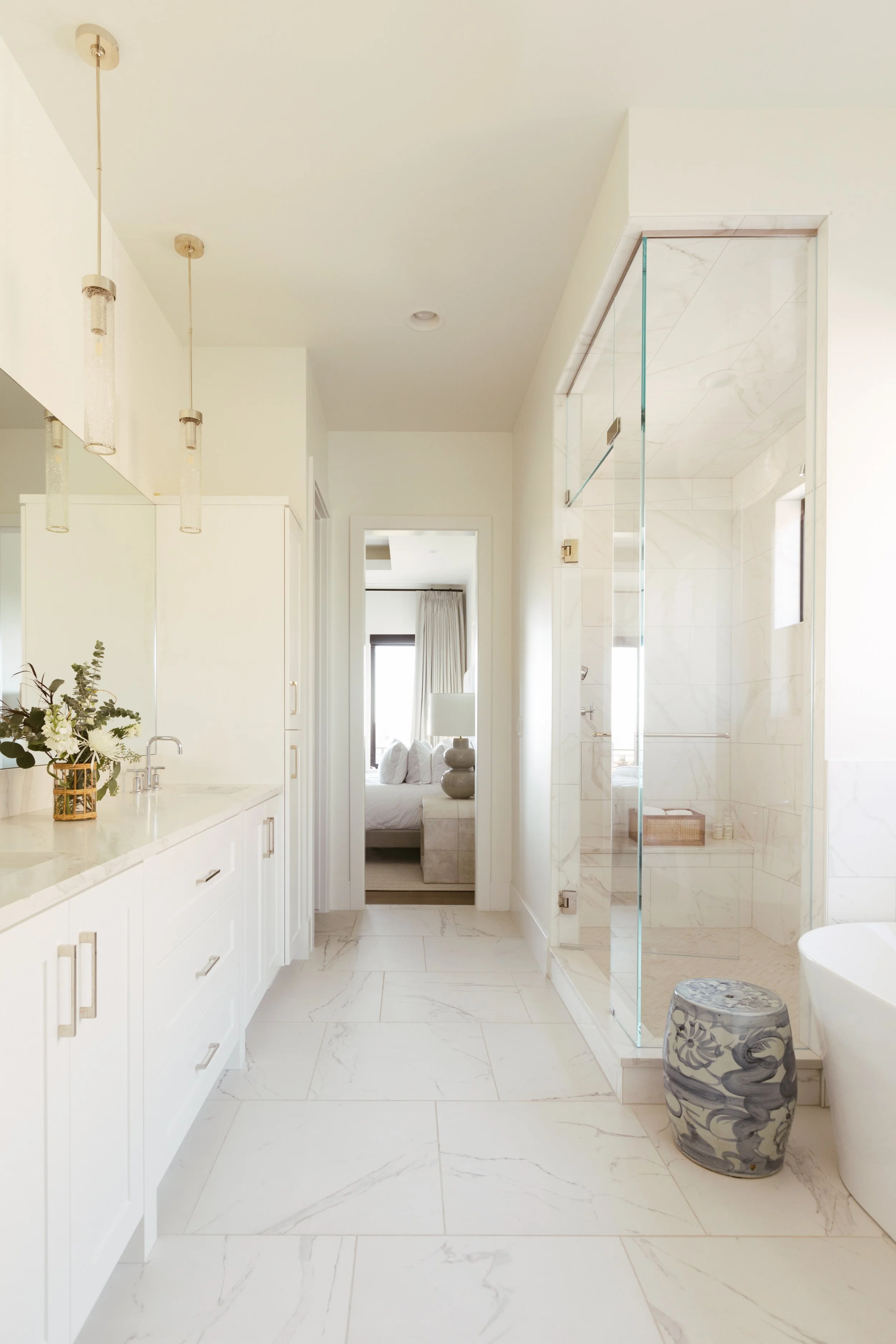 Modern bright bathroom with white cabinetry, marble flooring, and a glass shower enclosure, view into a bedroom with a bed and window with curtains.