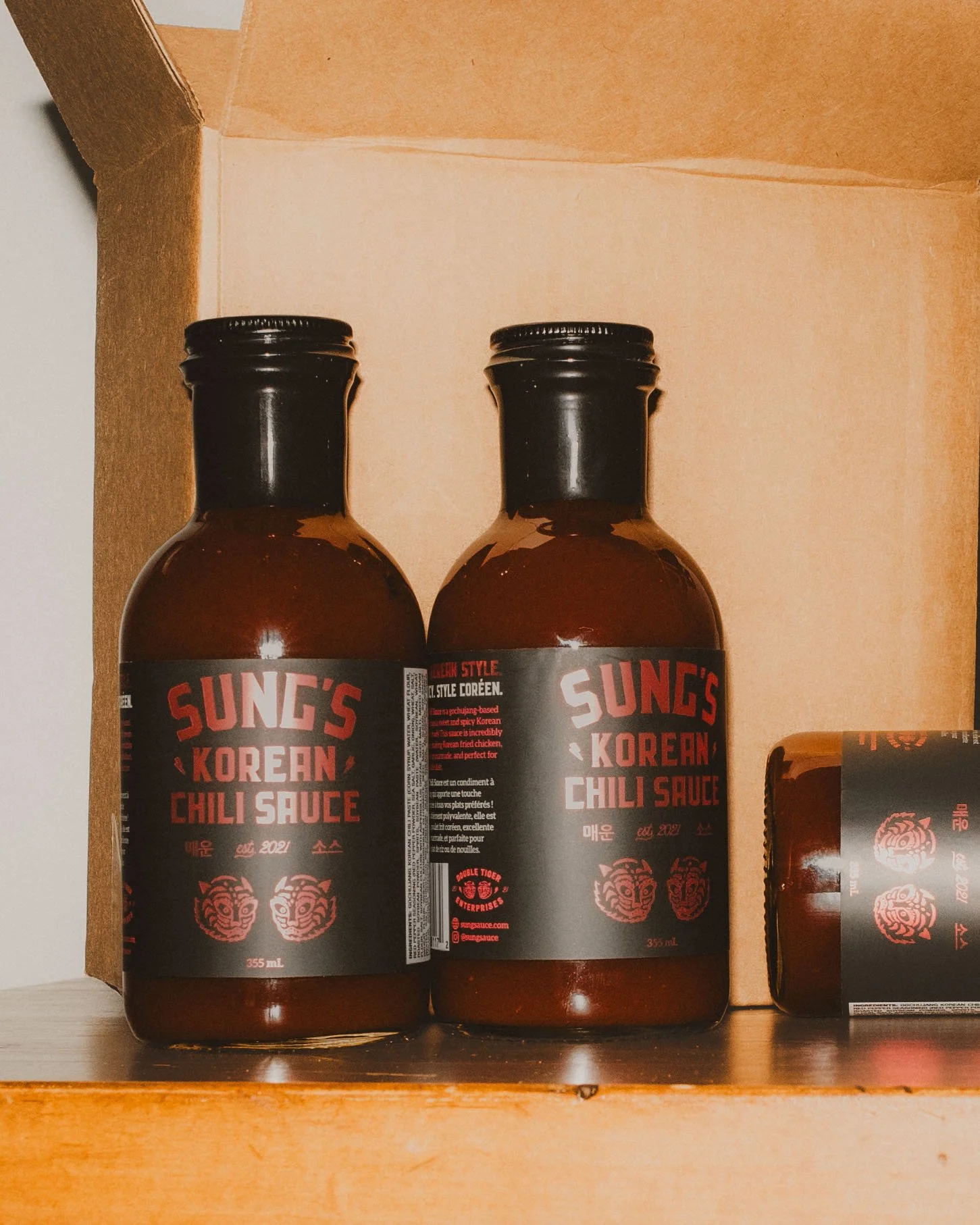 Two bottles of Sung's Korean Chili Sauce on a shelf with a cardboard box in the background.