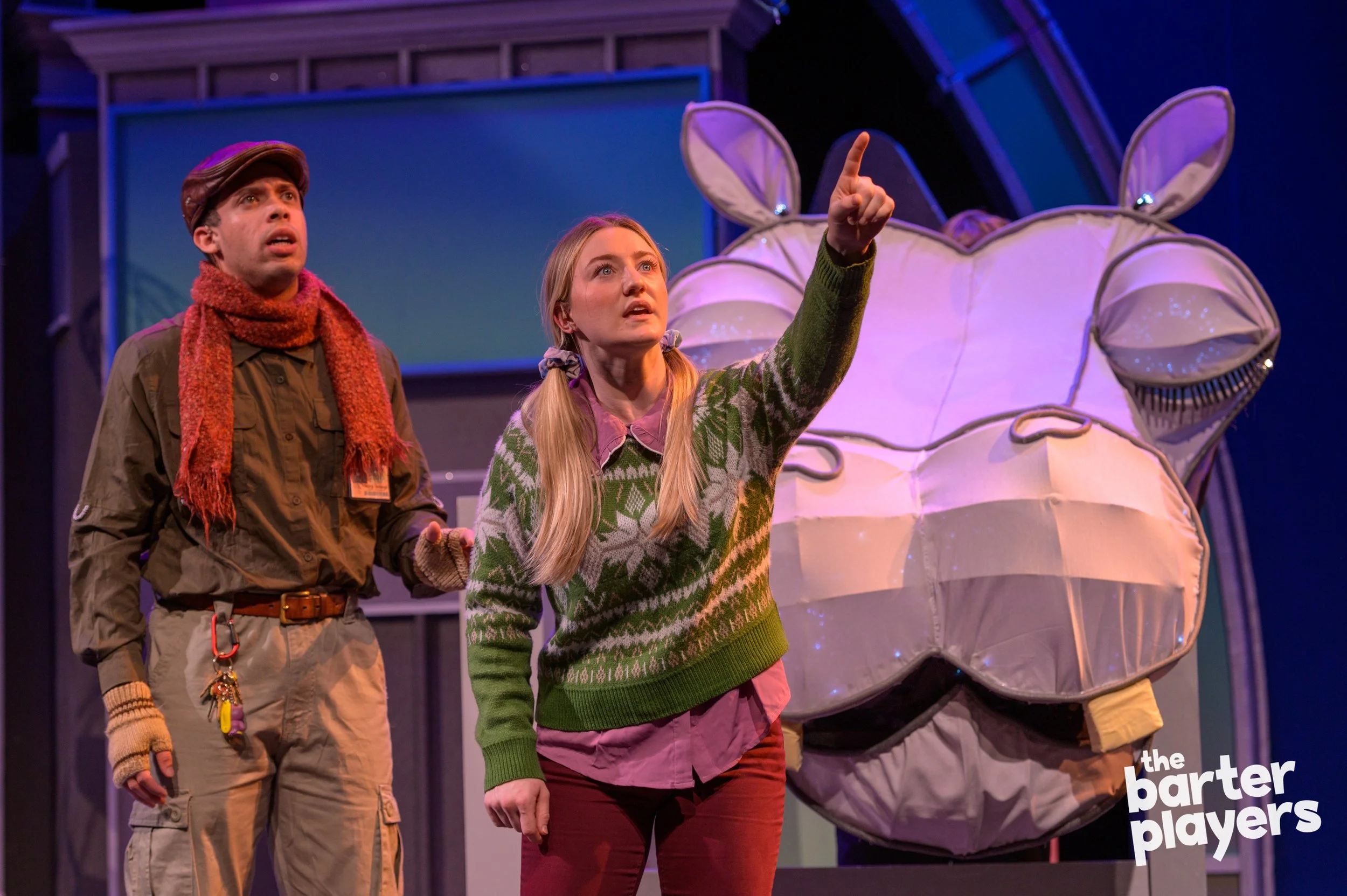 Two actors on stage, a man wearing a brown hat, scarf, and gloves, and a woman with long blonde hair in a green and pink patterned sweater, pointing towards something off-stage, with a large stylized pig face prop in the background, and the logo 'the barter players' in the bottom right corner.