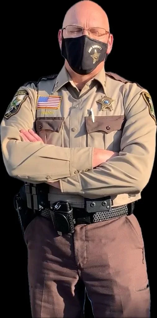 A uniformed police officer with a badge, American flag patch, and name tag that reads 'Nelson,' wearing a black face mask and glasses, stands with arms crossed against a black background.