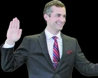 A man in a suit and tie smiling and raising his right hand in a wave.