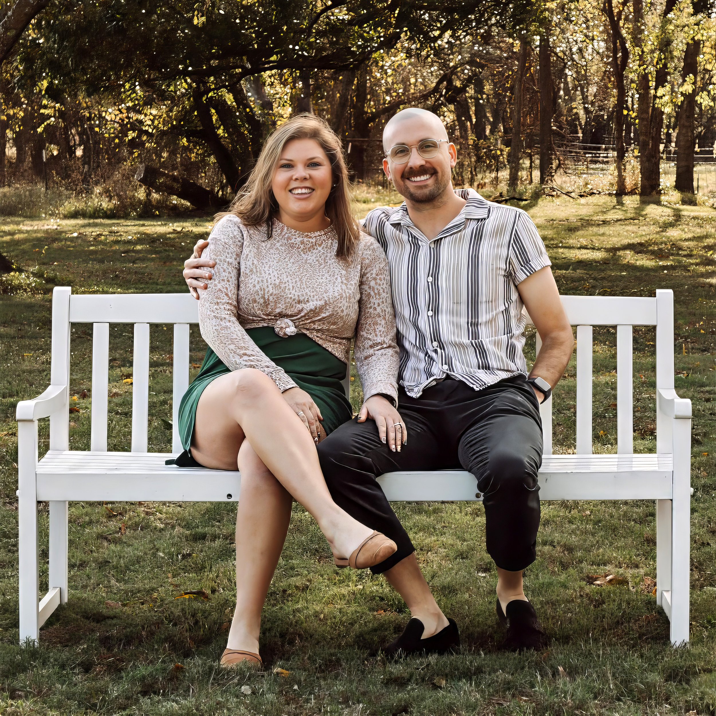 A smiling couple sitting closely together on a white bench outdoors in a park with trees and grass in the background.