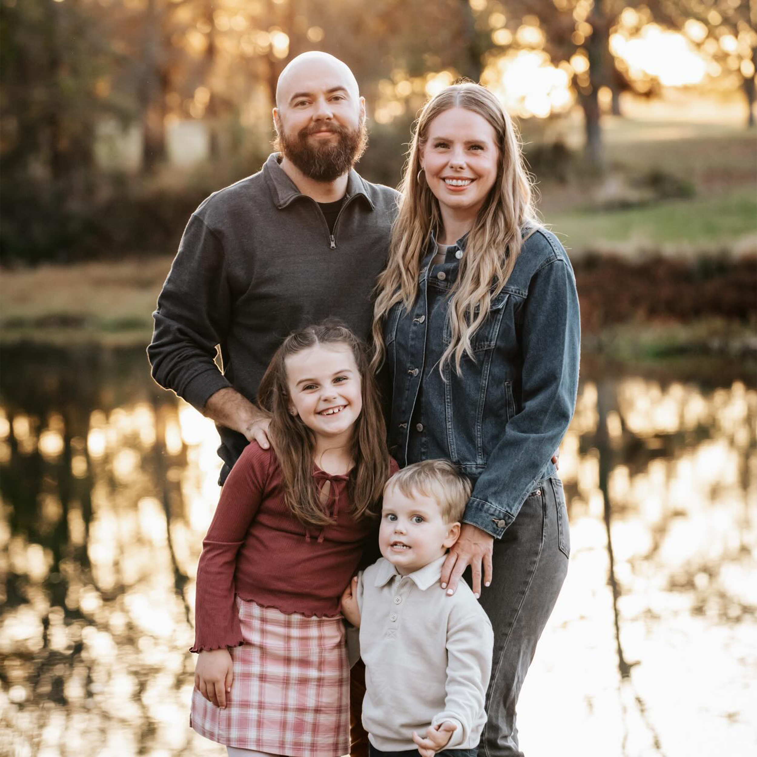 A family of five standing outdoors near a lake at sunset, smiling at the camera. The group includes a bald man with a beard, a blonde woman, a girl with brown hair in a red top and plaid skirt, and a young boy in a white sweater.