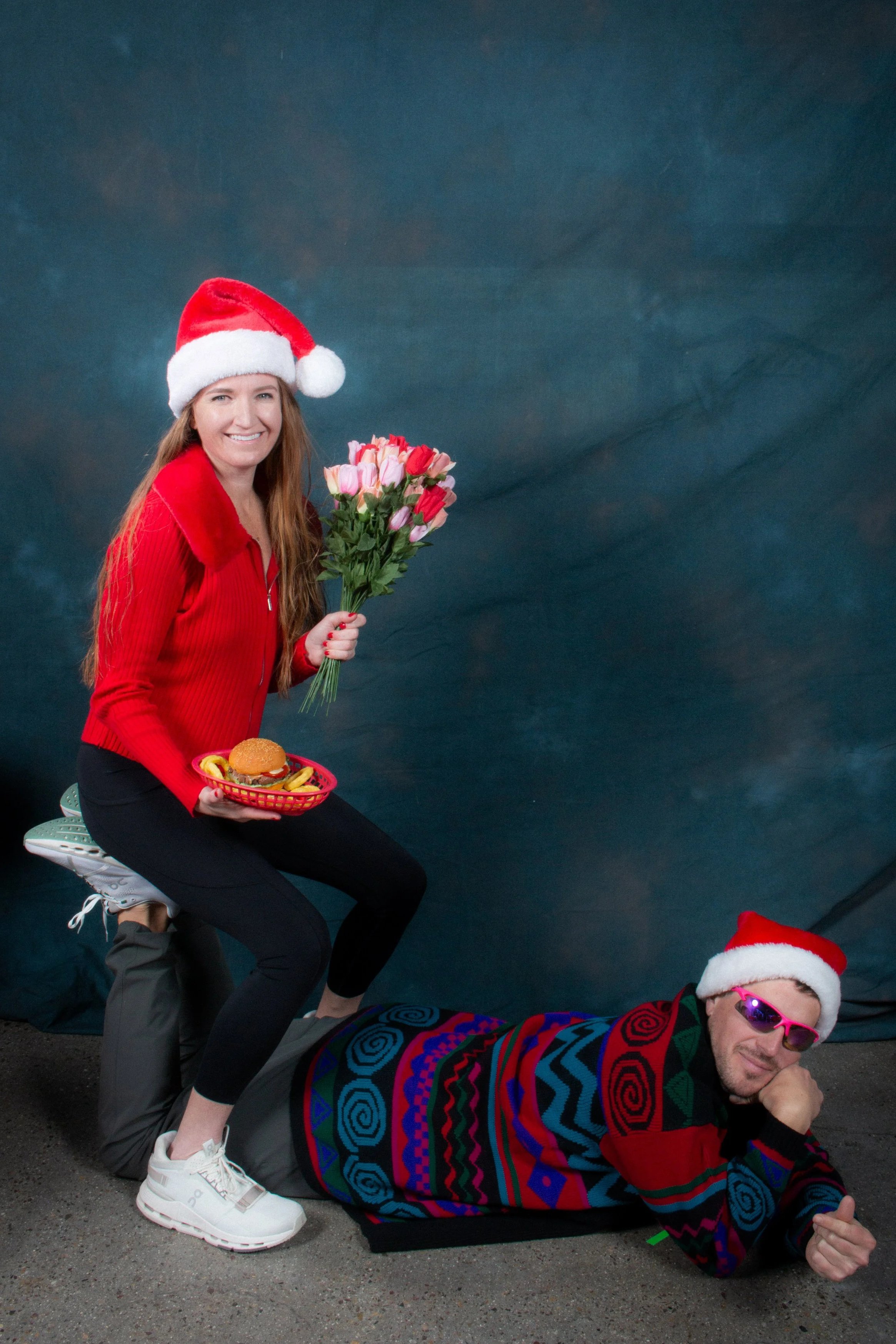 Three people dressed in Christmas attire, one woman wearing a Santa hat and holding a bouquet of flowers, another person wearing sunglasses and a Santa hat lying on the ground, and a third person in colorful clothing and a Santa hat lying on top of t