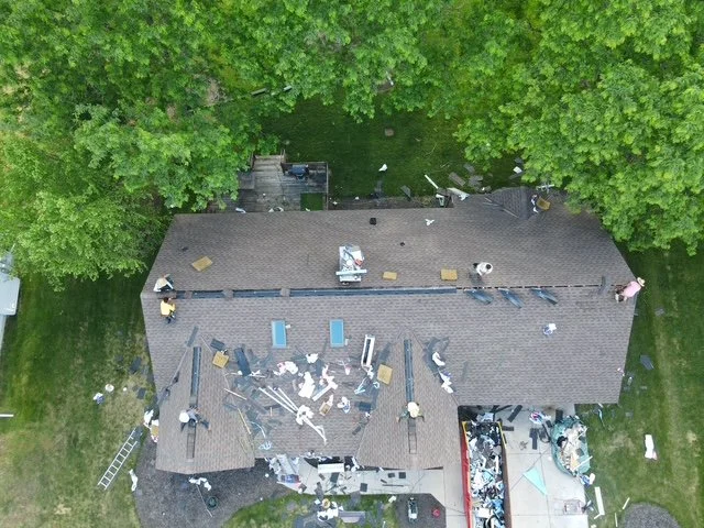 Aerial view of a house with a brown roof, showing signs of repair work. Debris, tools, and materials are scattered around, and workers are visible on the roof and around the property. Green trees surround the house.