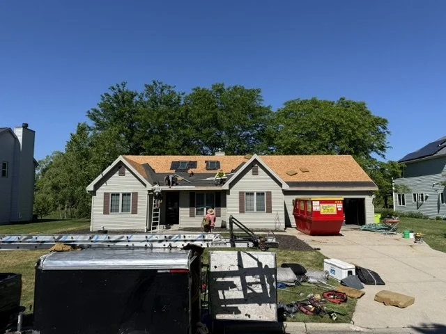 A house with a new roof under construction, a large red dumpster, and various tools and equipment in the driveway on a sunny day.