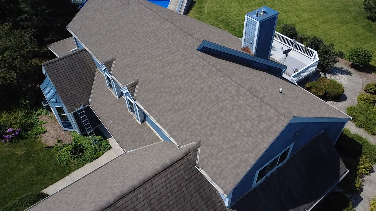 Aerial view of a house with a gray shingle roof, multiple dormer windows, and a rooftop deck with a satellite dish, surrounded by green lawns and bushes.