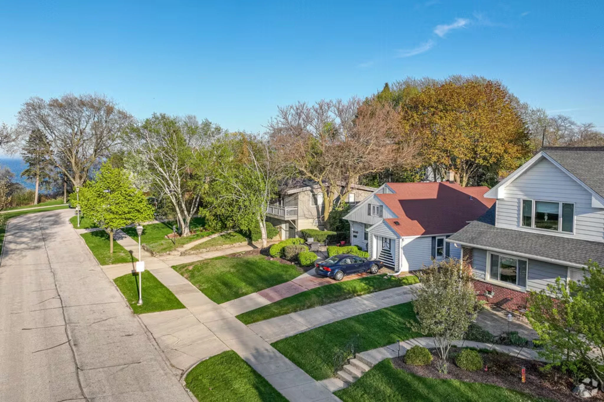 Aerial view of a suburban neighborhood featuring a white house with a red roof, a driveway with a parked car, sidewalk, green lawns, landscaped garden, trees, street lamps, and a curved street with clear blue sky and distant trees.