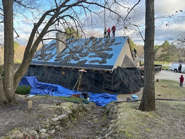 House with roof damage covered by black tarp and blue tarp, with workers inspecting the roof from the top on a cloudy day.