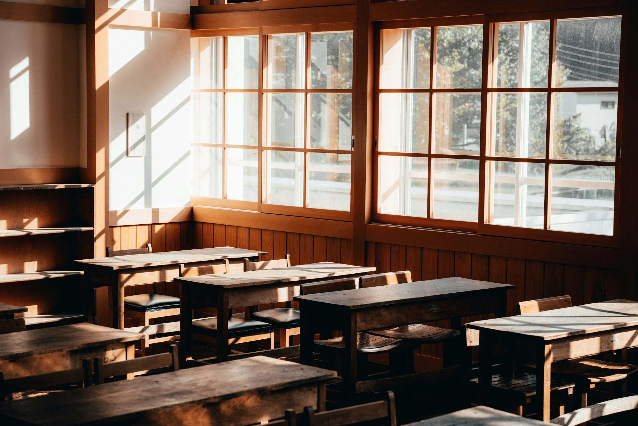 Sunlight streaming into a cozy classroom with empty wooden desks and chairs, large windows with wooden frames, and a wooden-paneled interior.