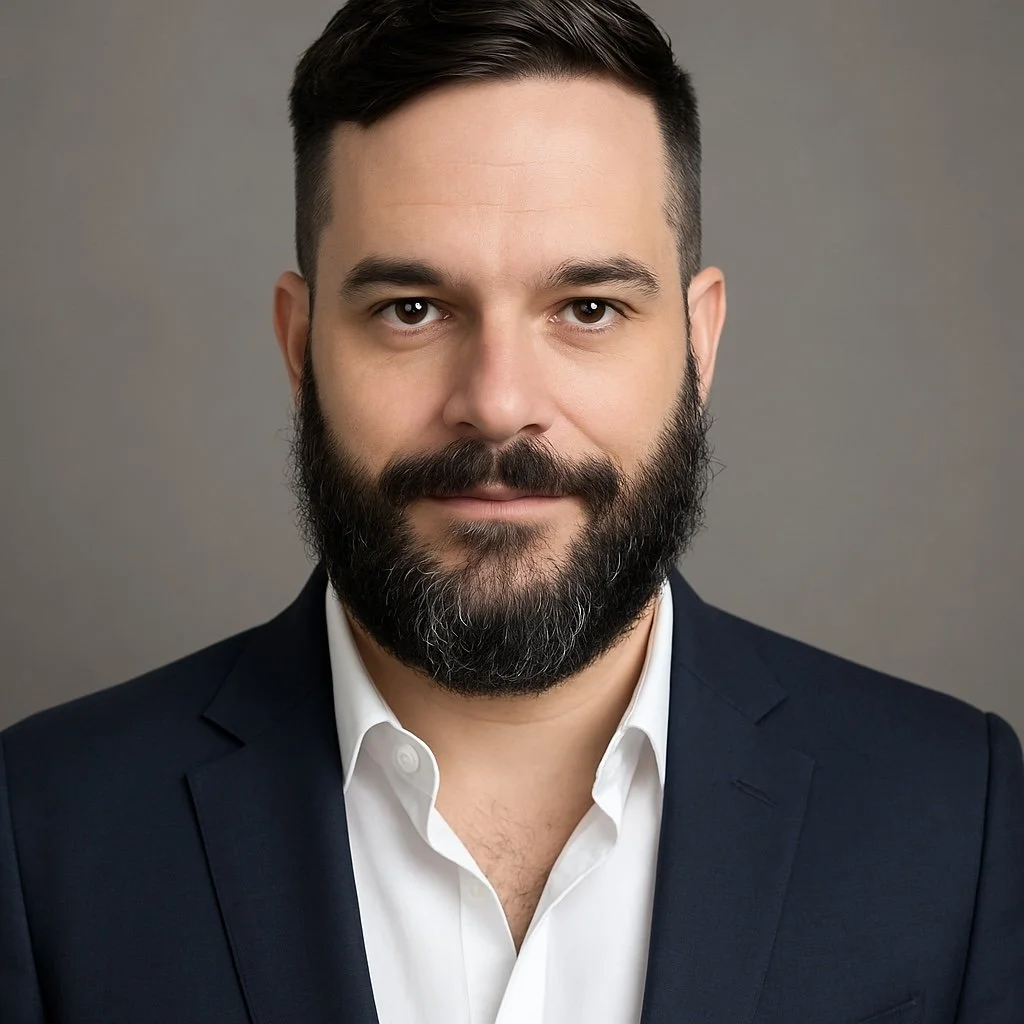 Headshot of a man with a beard, wearing a navy blazer and white shirt, against a gray background.