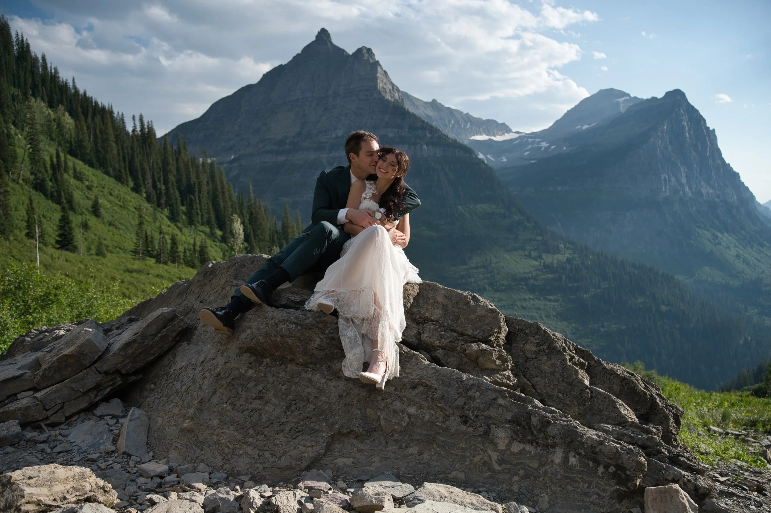 A couple dressed in wedding attire sitting on a large rock in a mountainous landscape with lush green trees and peaks in the background.