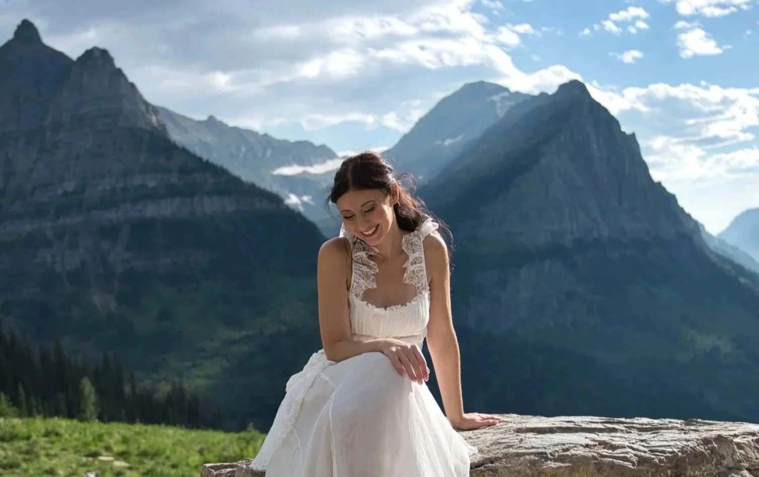 A woman in a white dress sitting on a rock outdoors, smiling, with mountains in the background under a partly cloudy sky.