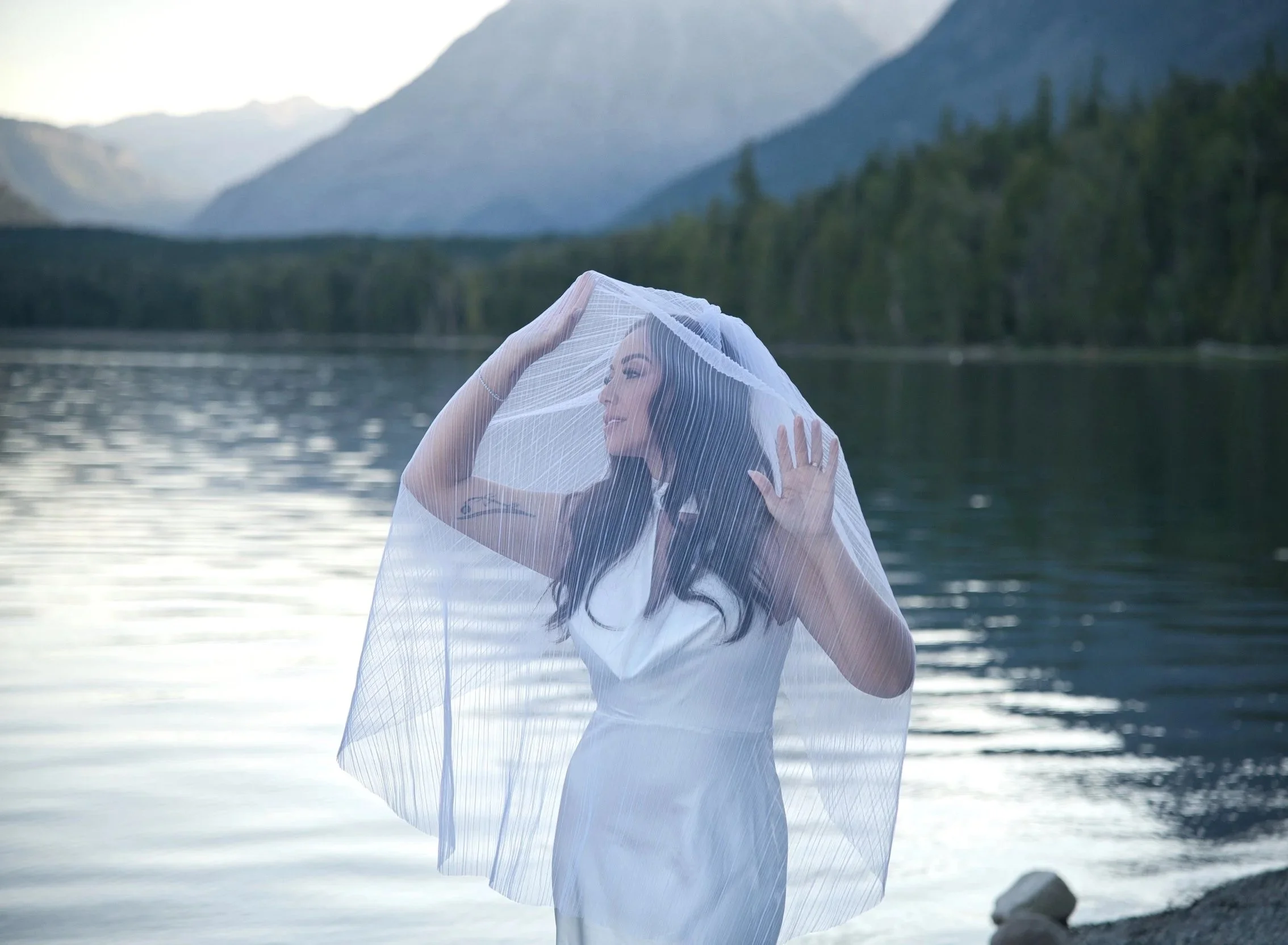 A woman with long dark hair standing by a lake with mountains in the background, holding a sheer white fabric over her head and shoulders.