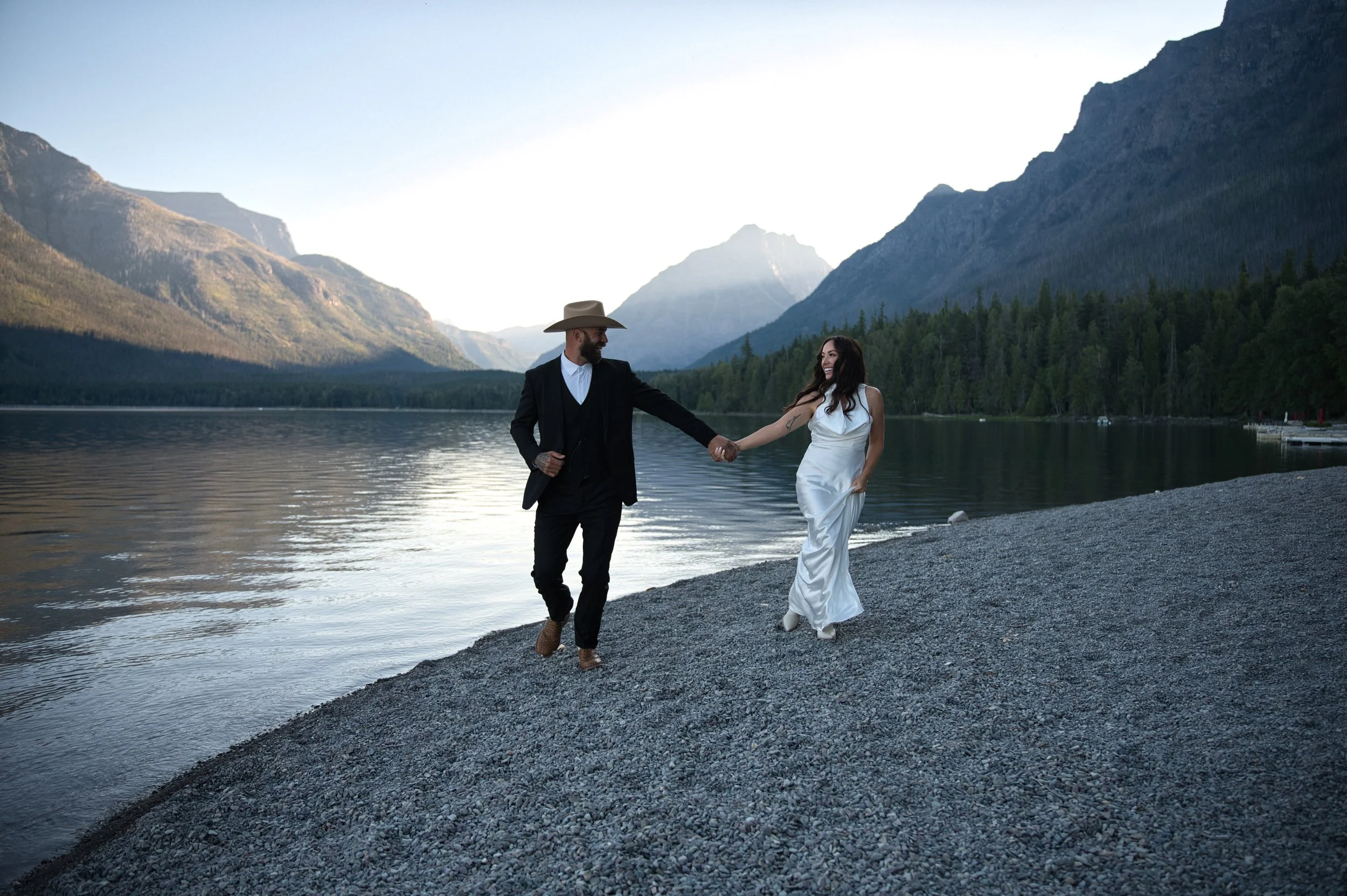 A man and woman holding hands and walking along a pebble beach near a lake with mountains and trees in the background. The man is wearing a black suit with a white shirt and a wide-brimmed hat, and the woman is in a white dress.