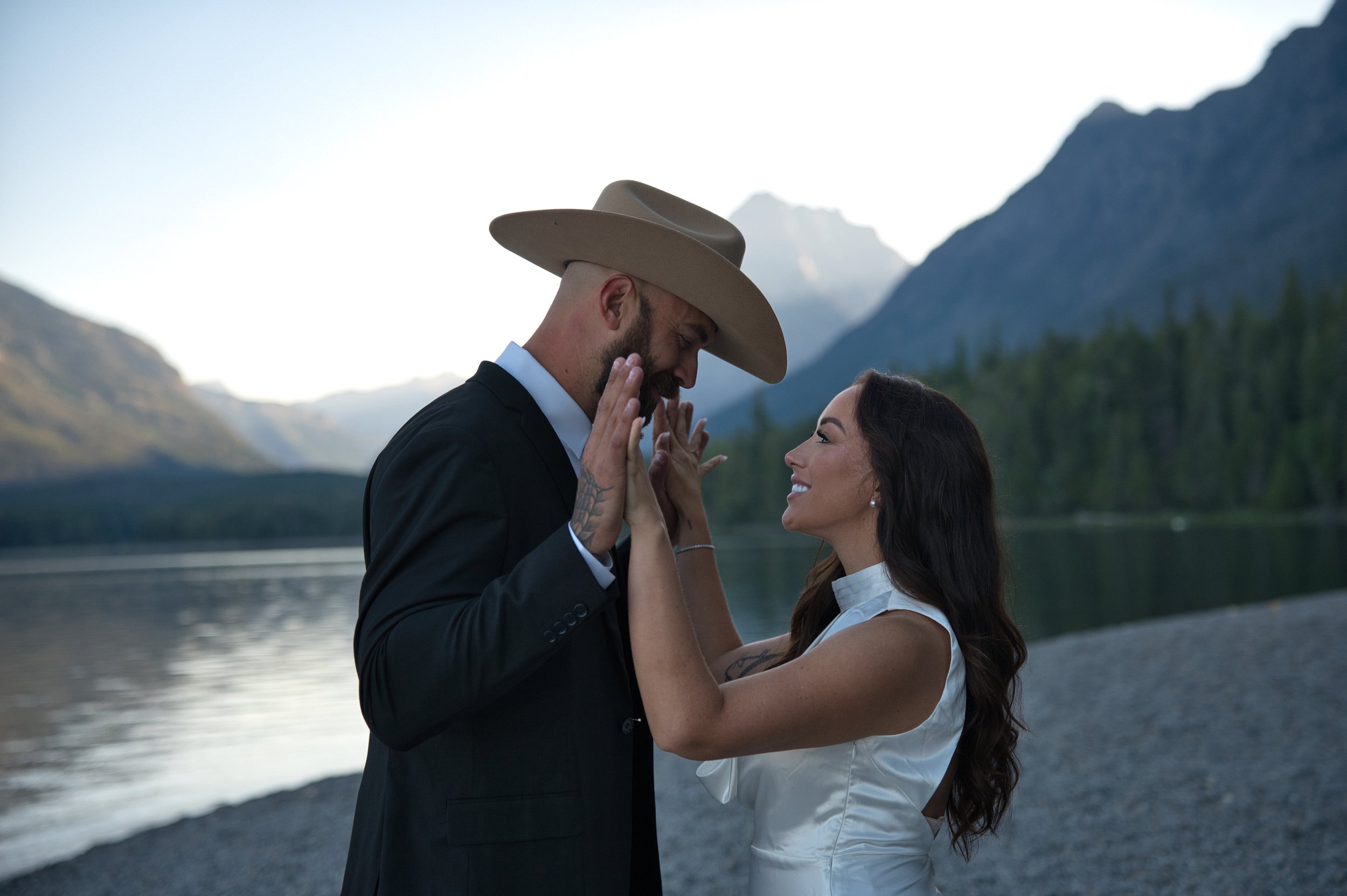 A couple standing by a lake with mountains in the background, smiling and touching each other's faces, dressed in formal and casual attire.