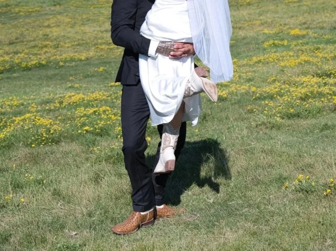 A groom holding a bride in a grassy field with yellow flowers, with the bride lifting one leg. The groom is wearing a suit and the bride is in a white dress with cowboy boots.
