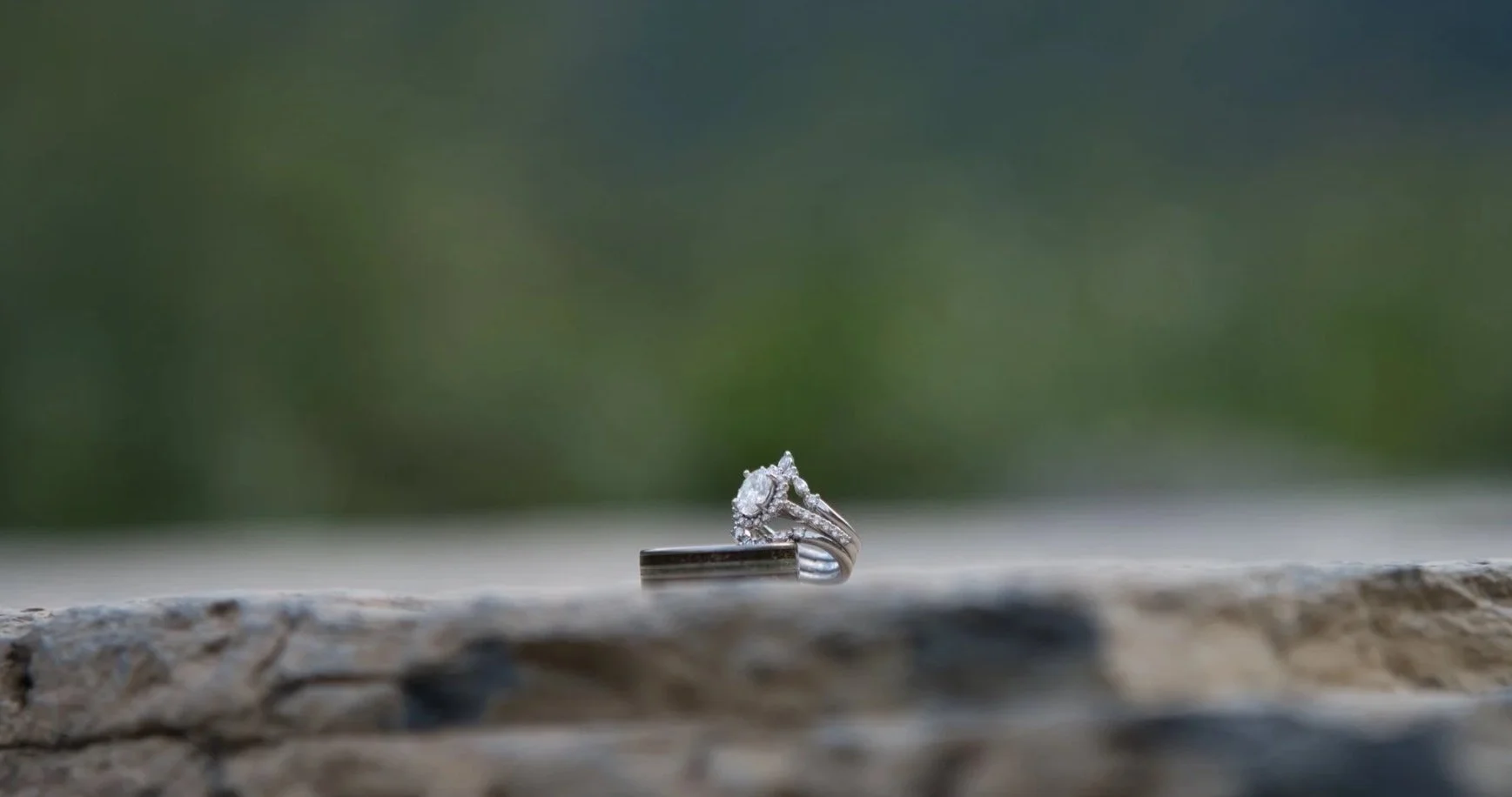 A diamond engagement ring with a large central stone surrounded by smaller diamonds, resting on a wooden surface with a blurred green background.