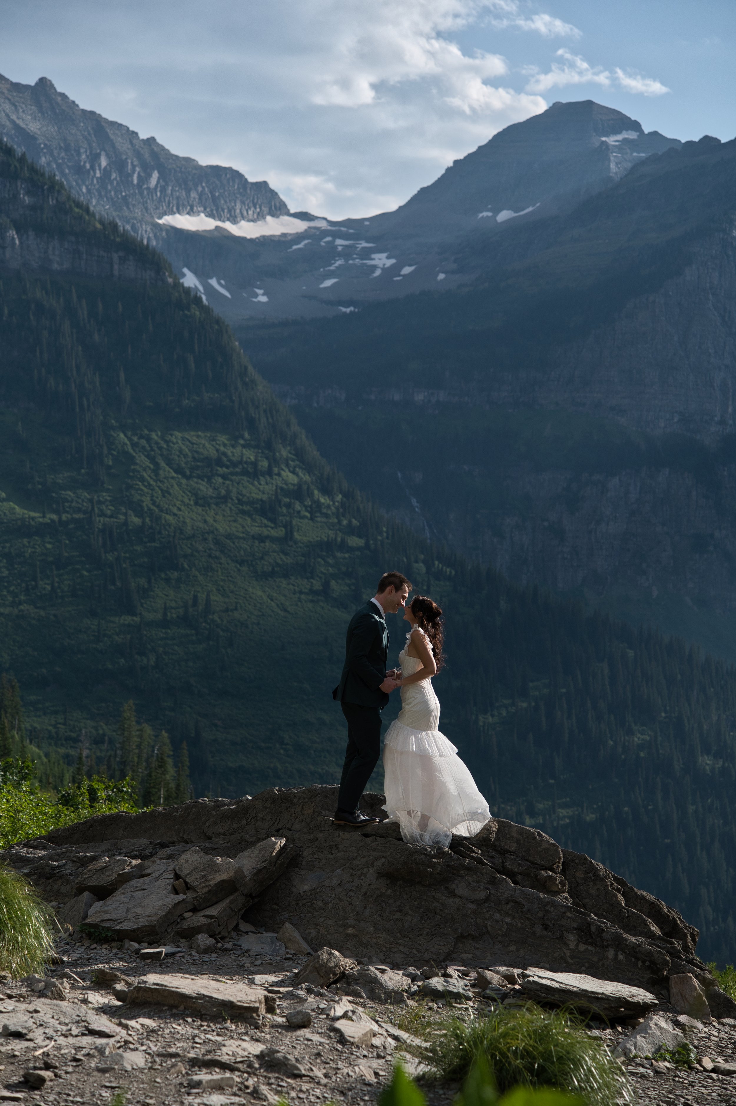 A bride and groom standing on a rocky outcrop in a mountainous landscape, with tall green mountains and snow patches in the background.