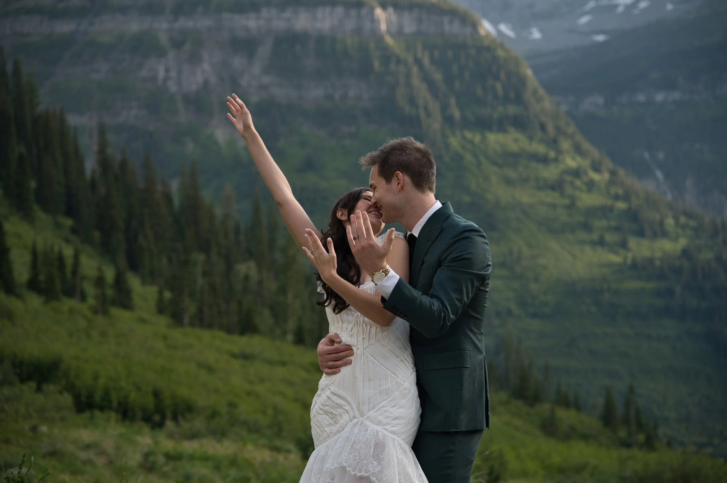 A couple in wedding attire sharing a romantic moment in a lush green mountain landscape. The woman is wearing a white dress, and the man is in a dark suit. They are embracing and smiling with their faces close together.