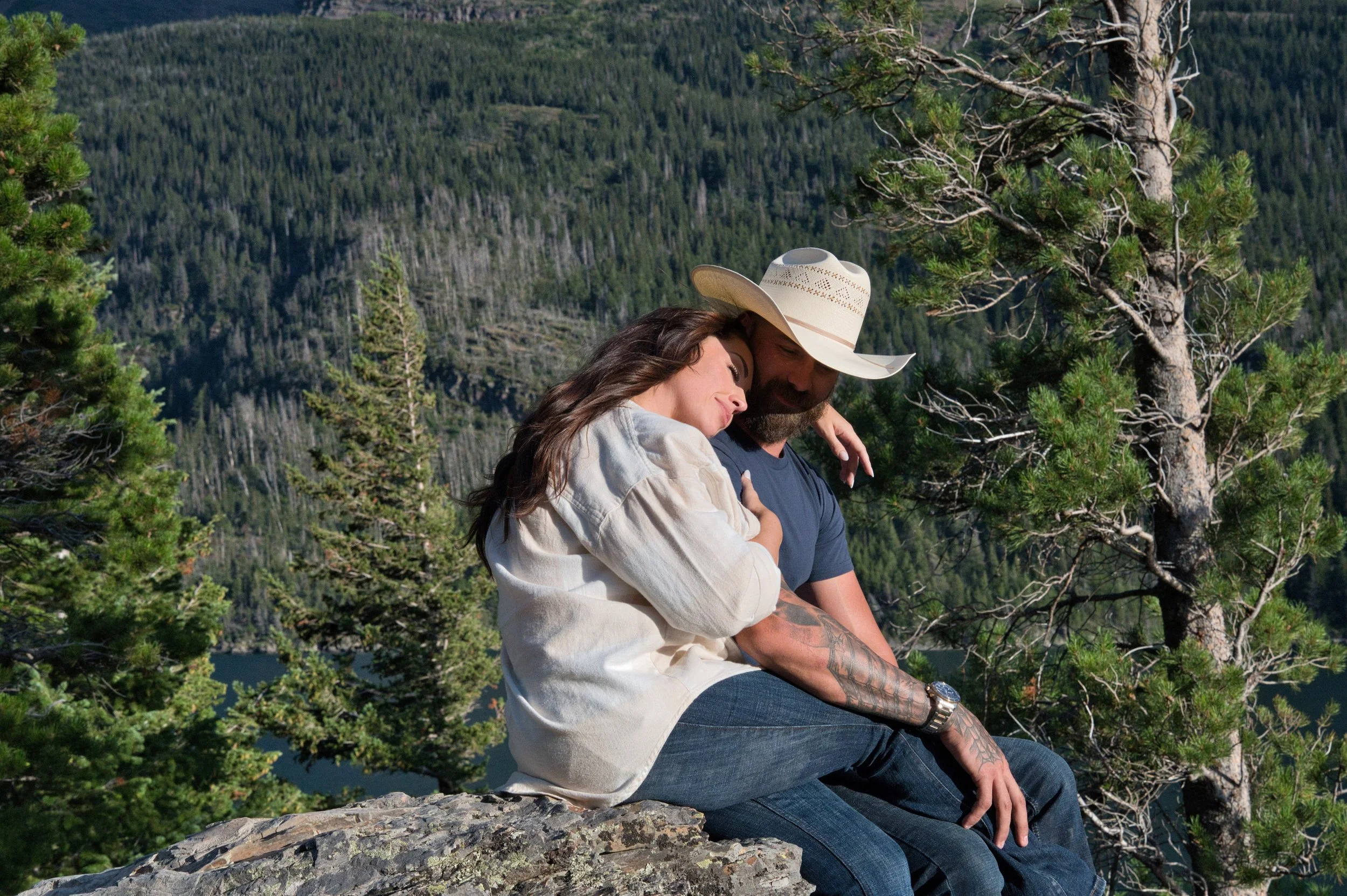 A couple sitting on a rock in a forested mountain area, with trees and mountains in the background, enjoying a moment together.