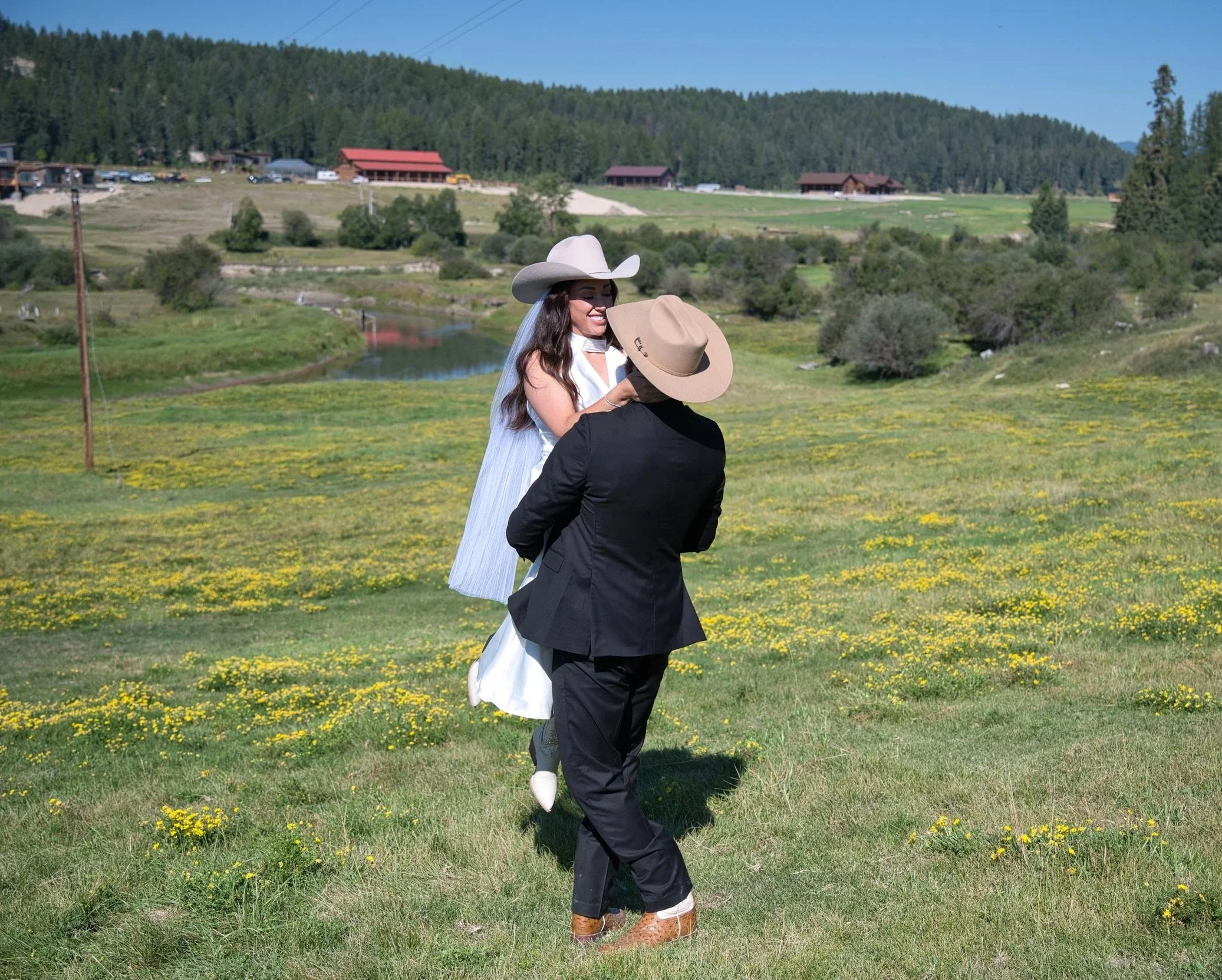 A couple dressed in cowboy attire standing in a grassy field with yellow flowers, holding each other and smiling. The woman wears a large white cowboy hat and a white dress, and the man wears a tan cowboy hat, black suit, and boots. In the background, there are hills, trees, houses, and a river.