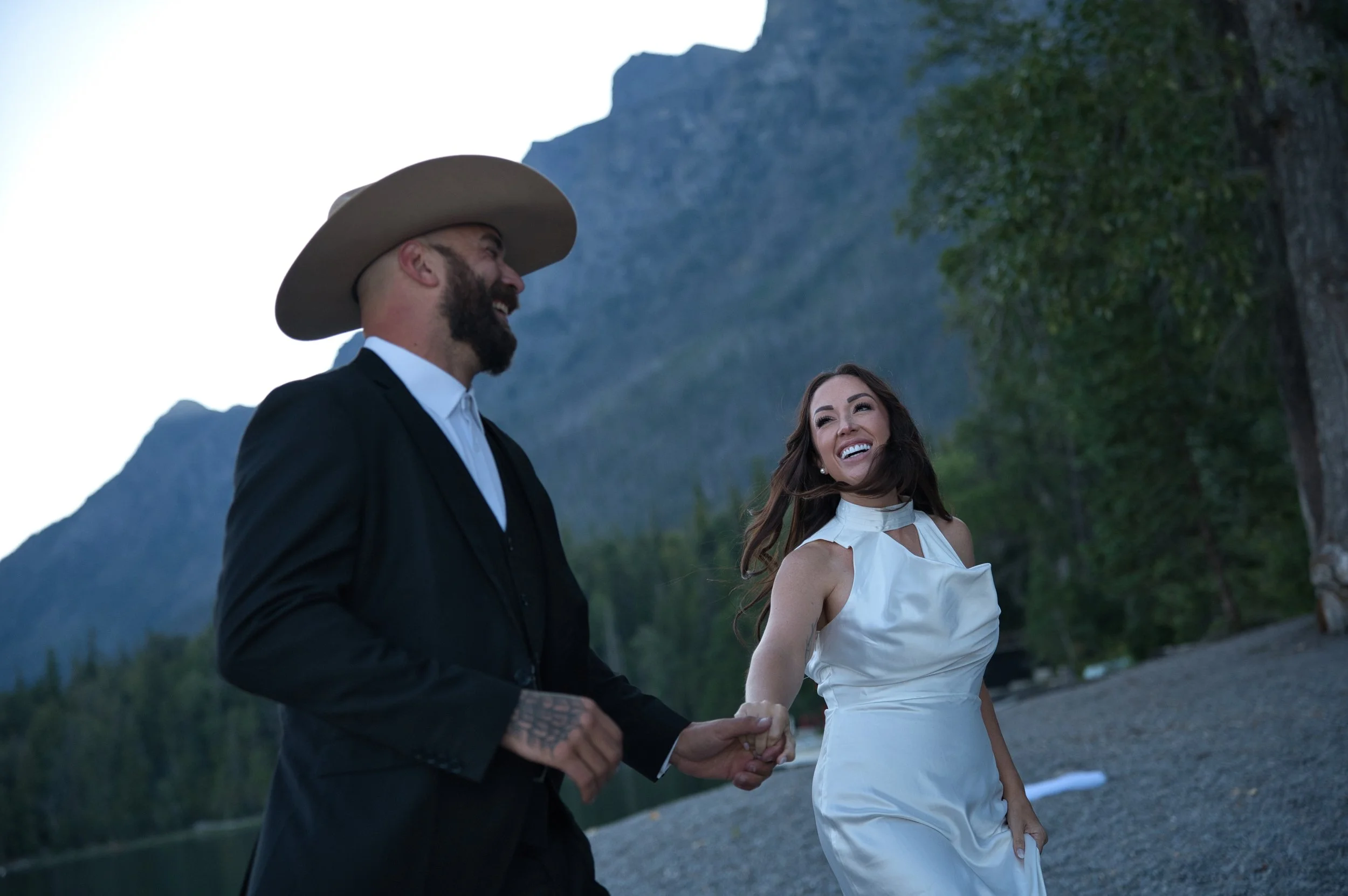 A smiling woman in a white dress holding hands with a man in a black suit and wide-brimmed hat, outdoors near a mountain and trees.