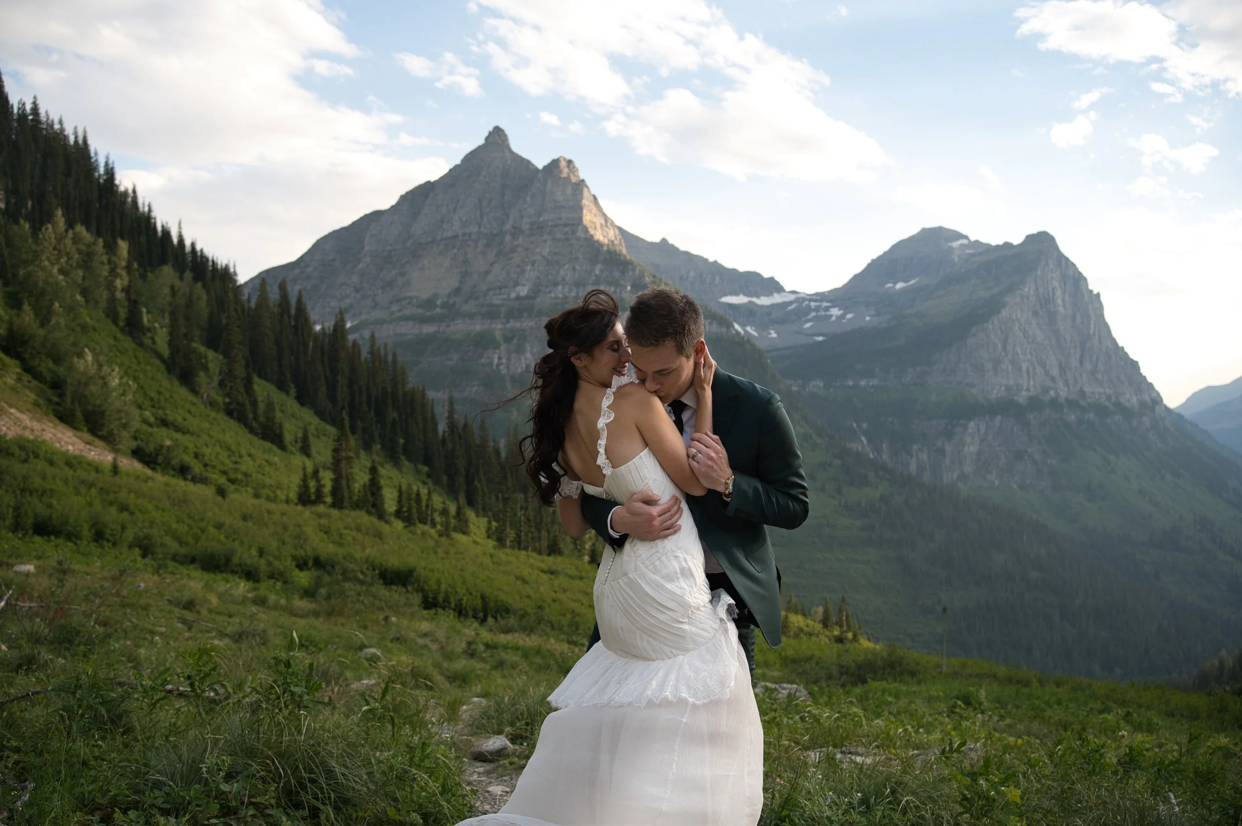 A couple in wedding attire embracing in a lush green mountain landscape with tall peaks in the background.